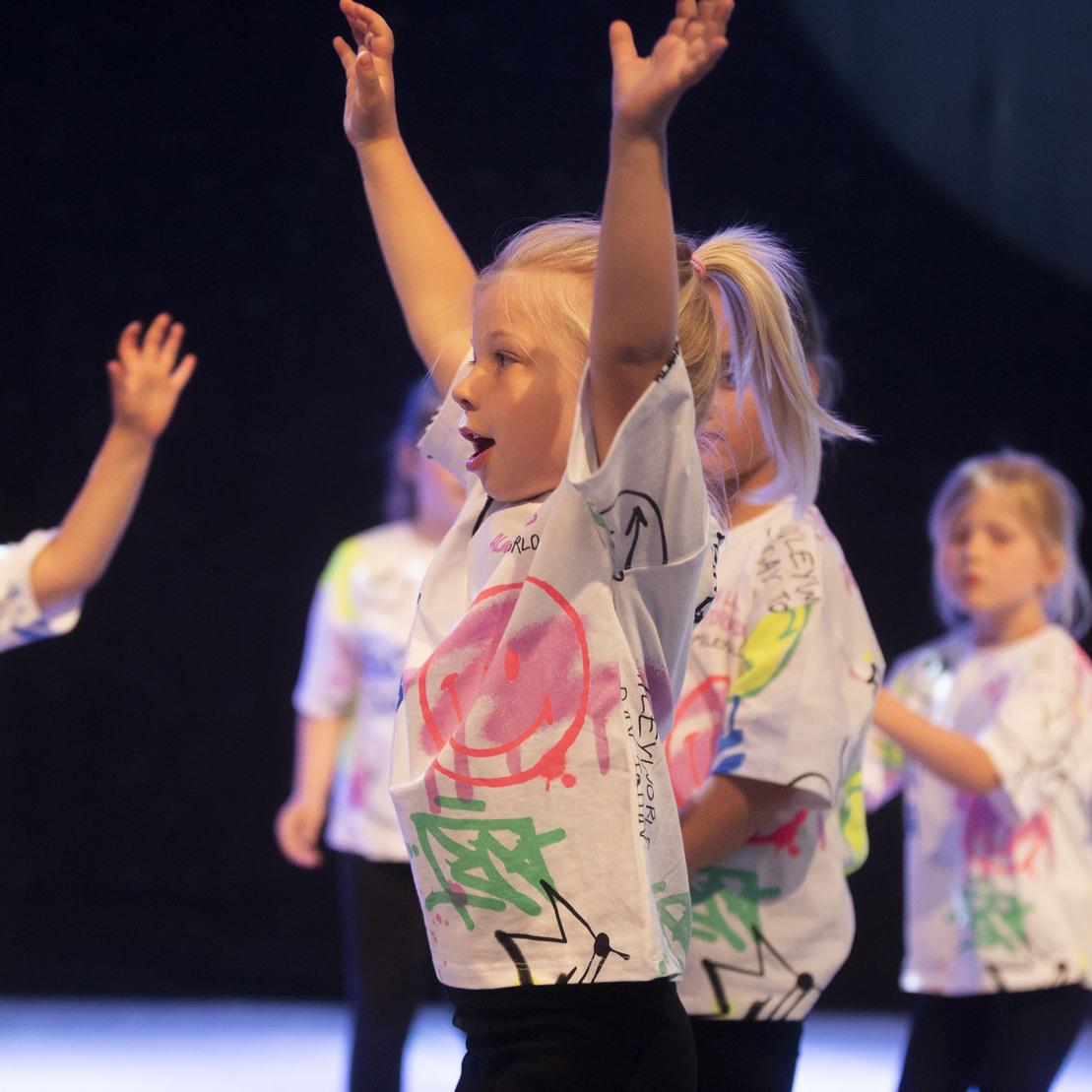 Gruppe von Kindern in bunten T-Shirts mit erhobenen H&auml;nden w&auml;hrend einer Tanzauff&uuml;hrung auf einer B&uuml;hne.