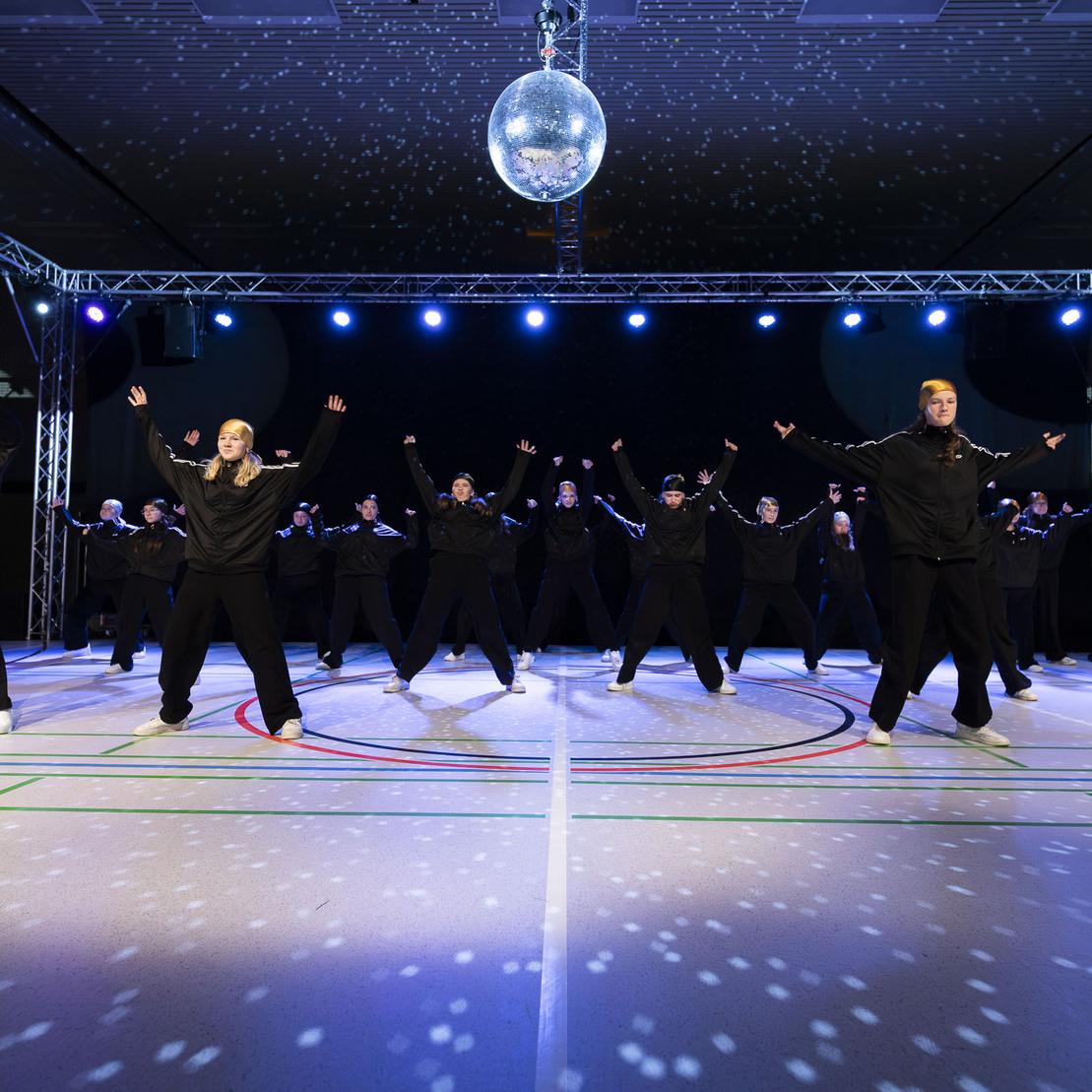 Tanzgruppe in schwarzen Outfits performt synchron unter einem Discokugellicht auf einer beleuchteten B&uuml;hne.
