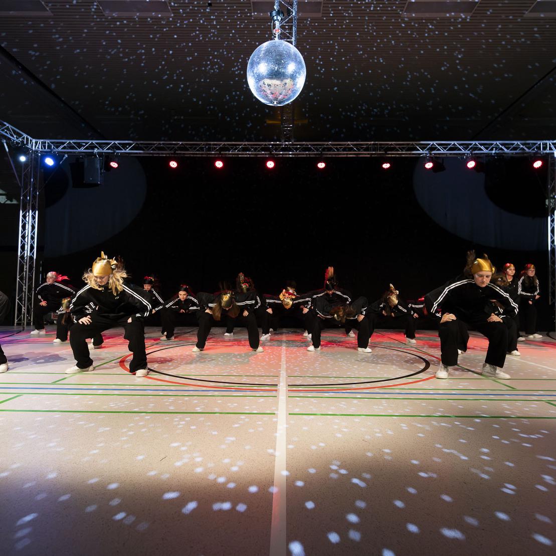 Tanzgruppe f&uuml;hrt synchronisierte Bewegungen unter einer Discokugel auf geflecktem Boden in einer Halle aus.