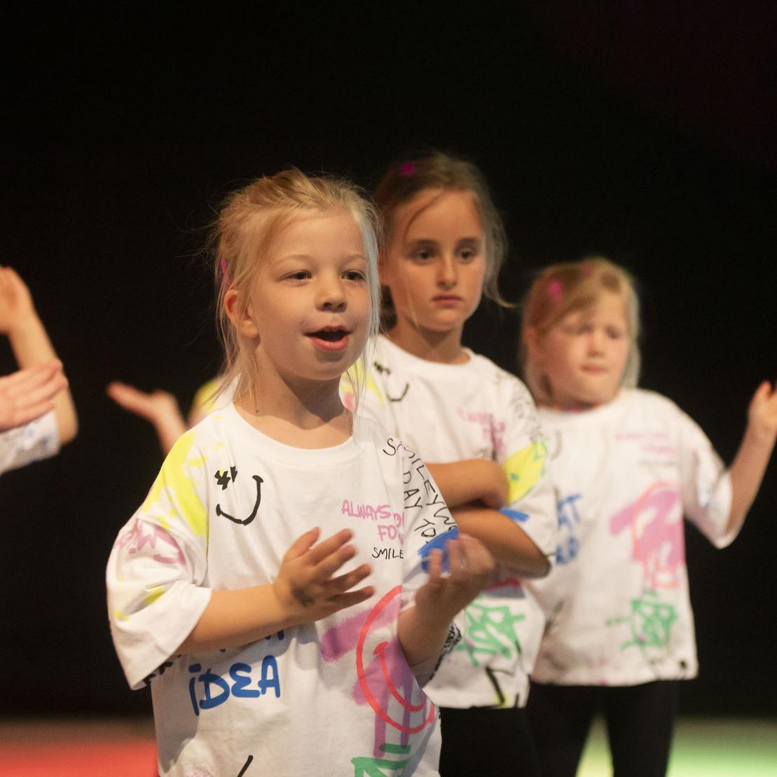 Sechs Kinder in bunten T-Shirts tanzen und lachen auf einer B&uuml;hne mit dimmtem Licht.