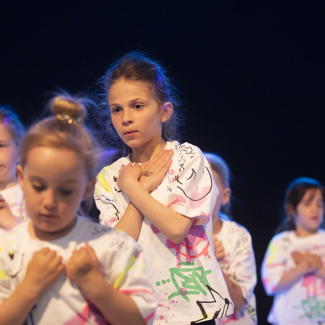 Tanzende Kinder in bunten T-Shirts stehen auf einer B&uuml;hne mit gedimmtem Licht und dr&uuml;cken ihre H&auml;nde ans Herz.