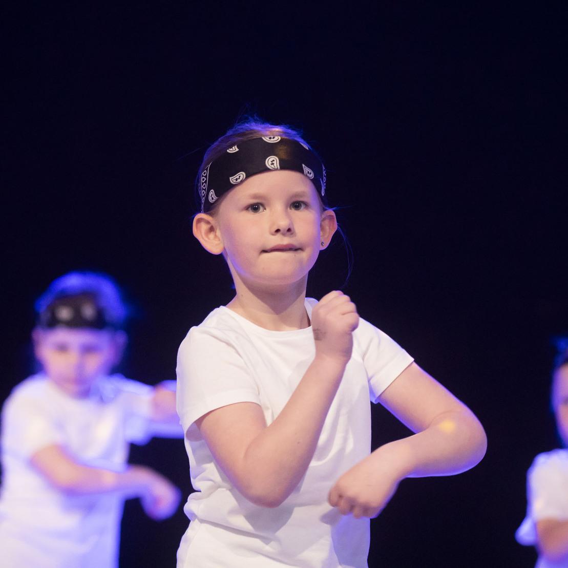 M&auml;dchen in wei&szlig;em T-Shirt mit Bandana tanzt auf einer B&uuml;hne, im Hintergrund weitere tanzende Kinder.