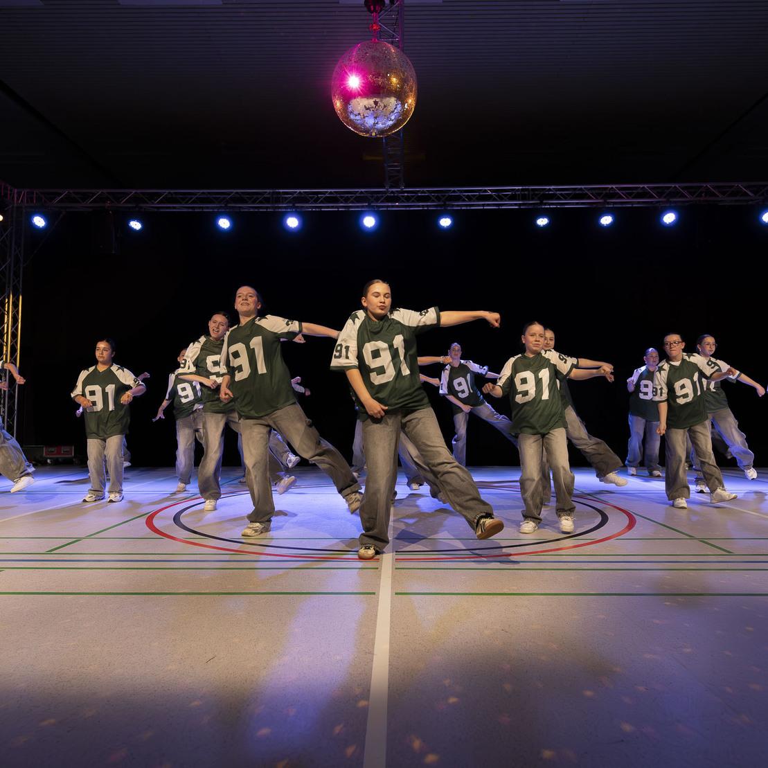 Tanzgruppe in gr&uuml;nen Shirts mit der Nummer 91 f&uuml;hrt eine synchronisierte Choreografie auf einer beleuchteten B&uuml;hne vor.