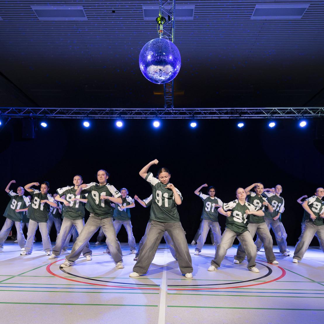 Tanzgruppe in gr&uuml;nen Trikots demonstriert synchron eine Choreografie unter einer Discokugel auf einer beleuchteten B&uuml;hne.