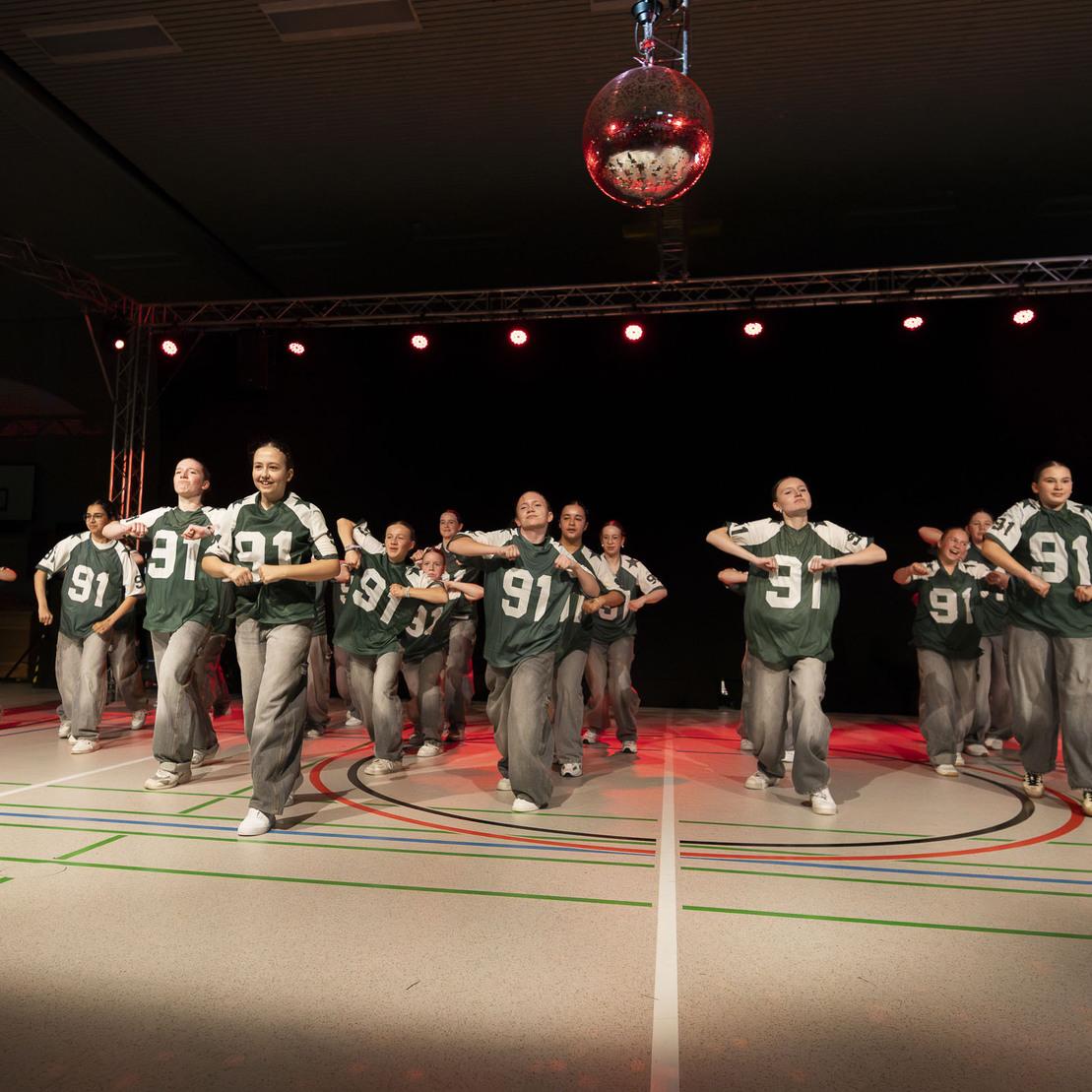 Tanzgruppe in gr&uuml;nen Trikots und grauen Hosen f&uuml;hrt eine Choreografie auf einer beleuchteten B&uuml;hne auf.