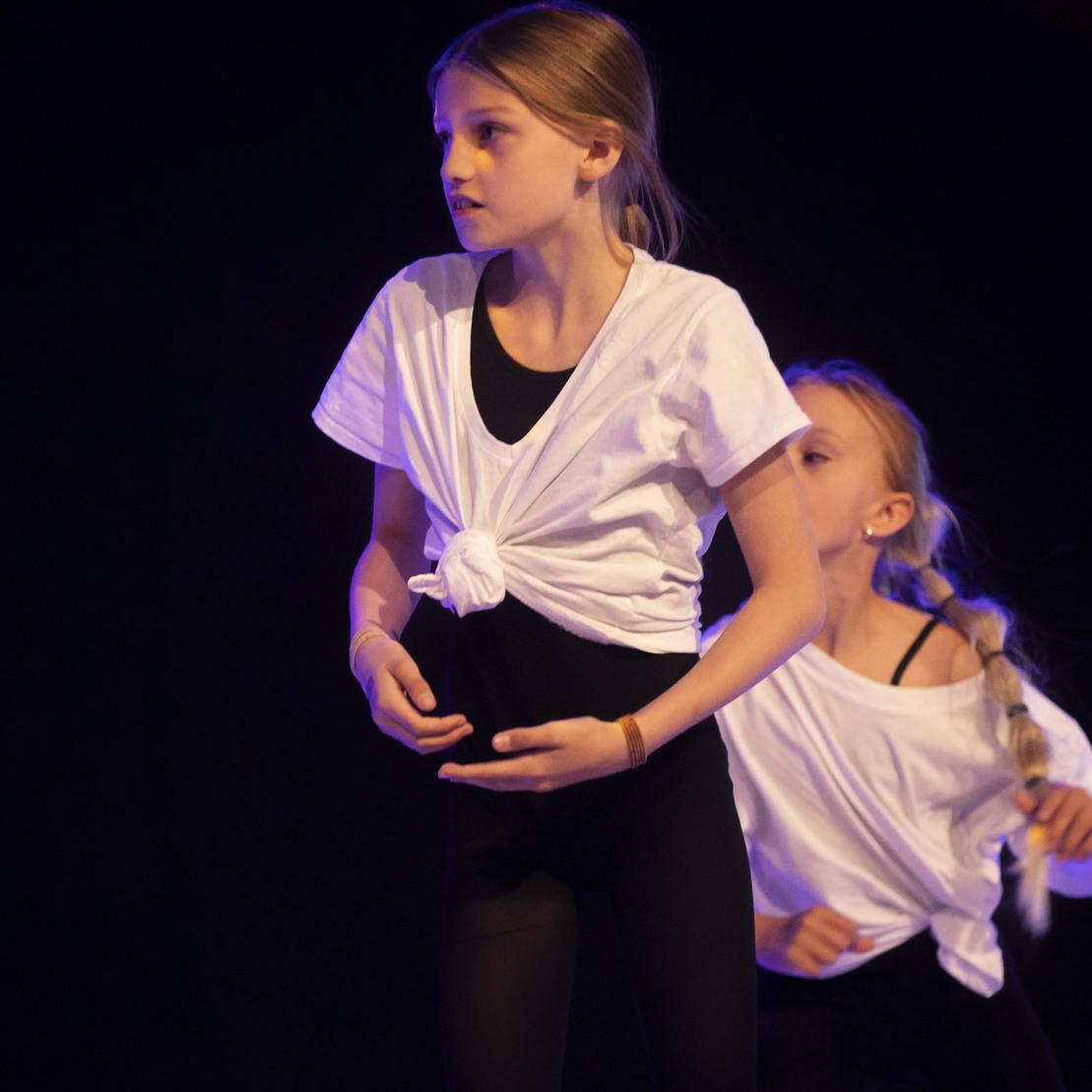 Zwei junge T&auml;nzerinnen in wei&szlig;en T-Shirts f&uuml;hren eine choreografierte Tanzbewegung auf dunkler B&uuml;hne aus.