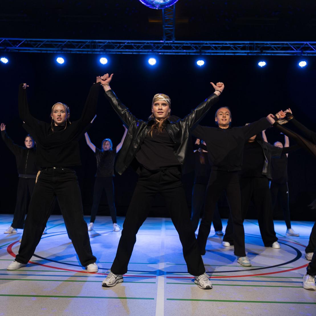 Tanzgruppe in schwarzen Outfits f&uuml;hrt eine synchronisierte Choreografie auf einer B&uuml;hne mit blauen Lichtern aus.