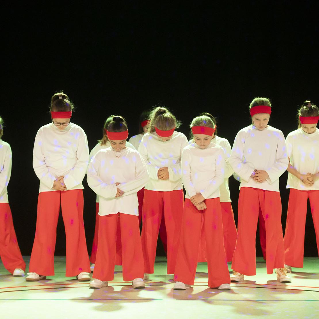 Gruppe von acht T&auml;nzerinnen in wei&szlig;en Oberteilen und roten Hosen, mit Kopfband, stehen auf einer B&uuml;hne in dekorativer Beleuchtung.