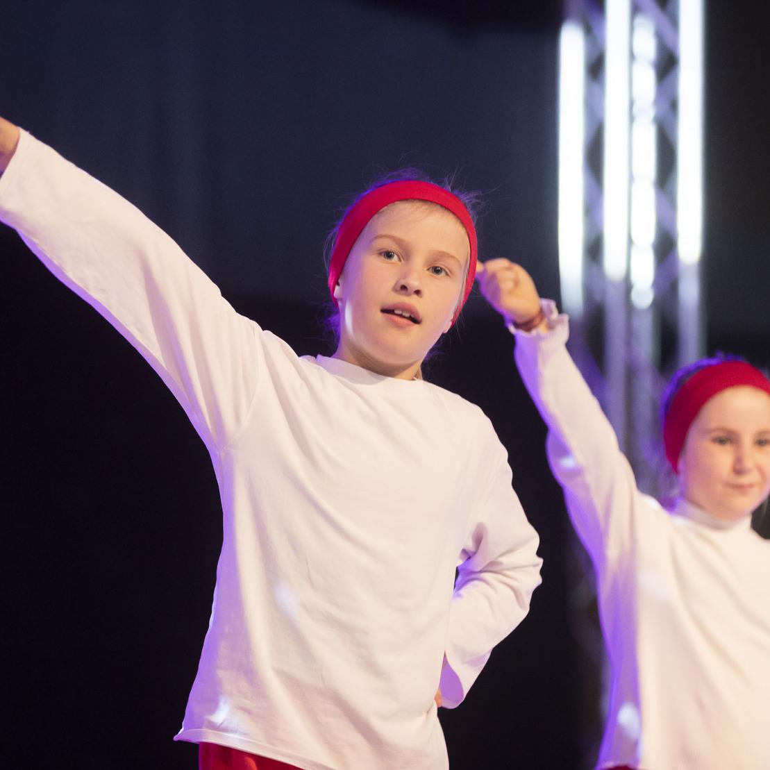 Zwei Kinder in wei&szlig;en Oberteilen und roten Haarb&auml;ndern f&uuml;hren eine Tanzroutine auf einer B&uuml;hne aus.