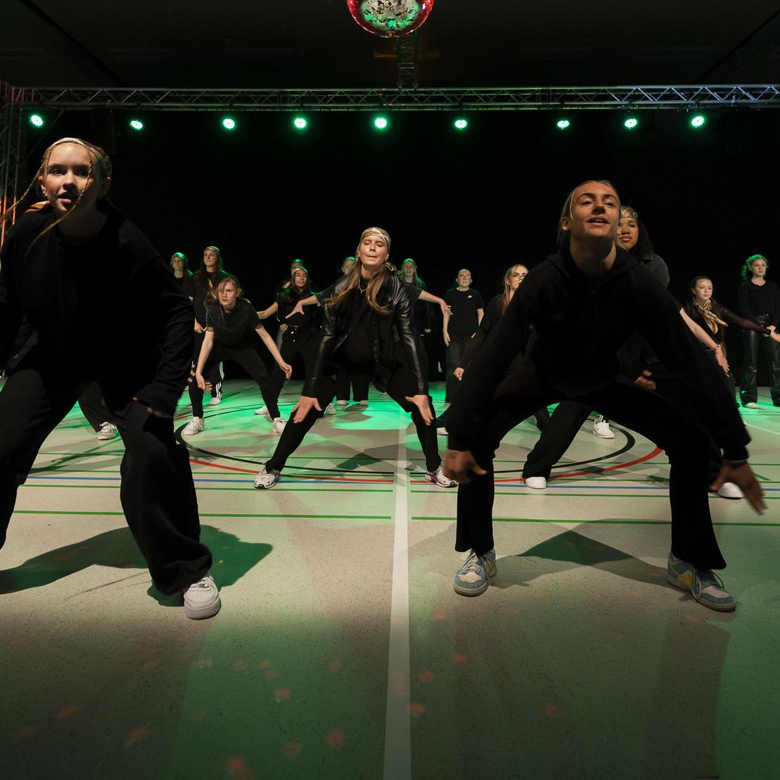 T&auml;nzerinnen in schwarzen Kost&uuml;men f&uuml;hren eine choreografierte Auff&uuml;hrung auf einer Sporthalle mit gr&uuml;nem Licht auf.