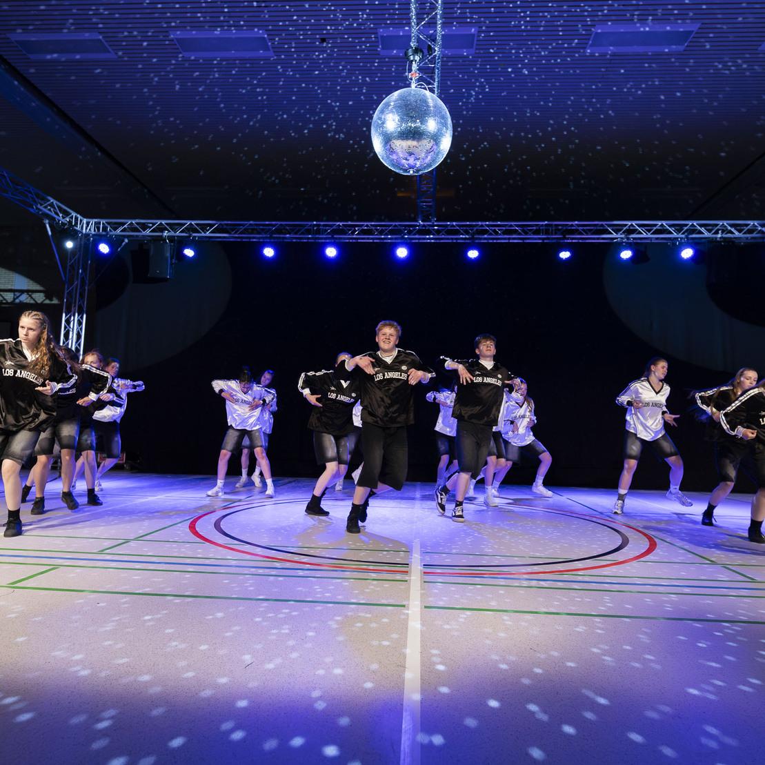 Tanzgruppe in sportlicher Kleidung f&uuml;hrt eine Choreografie unter bunten Lichtern und Discokugel auf einer Sportfl&auml;che vor.