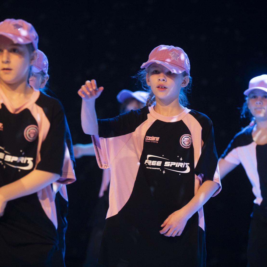 Gruppe von tanzen M&auml;dchen in Sportkleidung und Baseballcaps, die auf einer B&uuml;hne choreografierte Bewegungen ausf&uuml;hren.