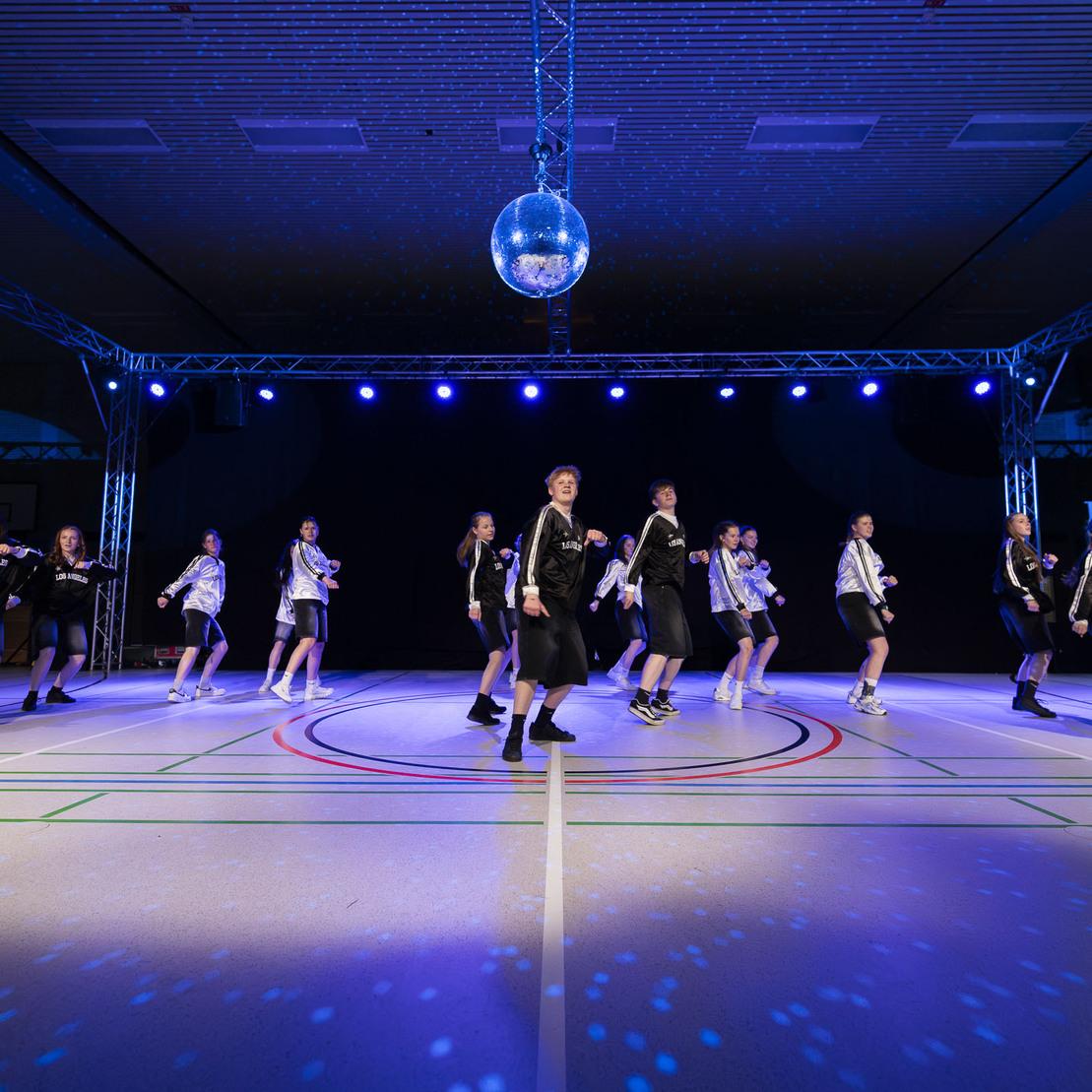 Tanzgruppe in Schwarz-Wei&szlig;-Kleidung performt auf einer Sportb&uuml;hne unter einem Disco ball und bunten Lichtern.