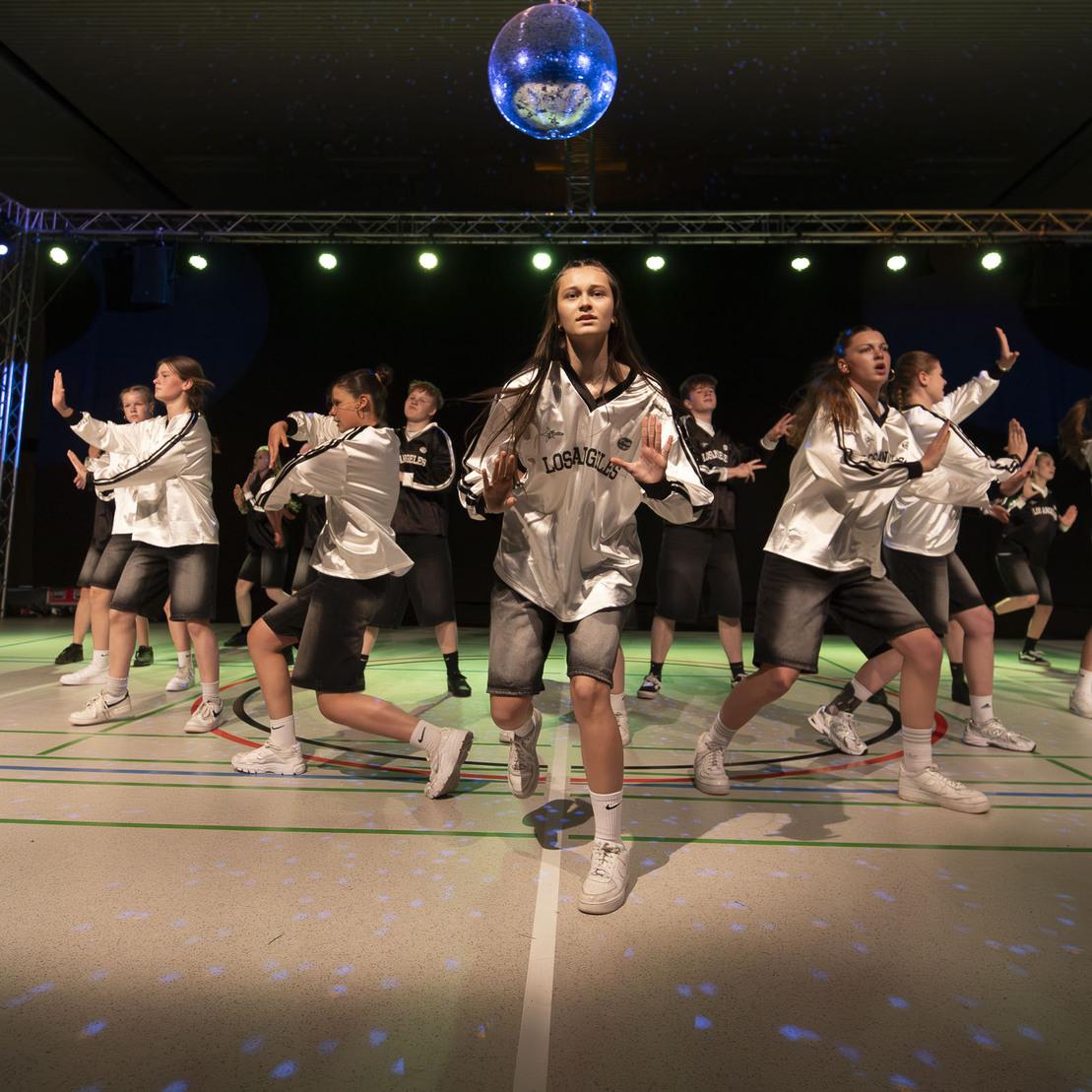 Tanzgruppe in schicken Outfits f&uuml;hrt eine Choreografie auf einer B&uuml;hne mit Discokugel und bunten Lichtern auf.