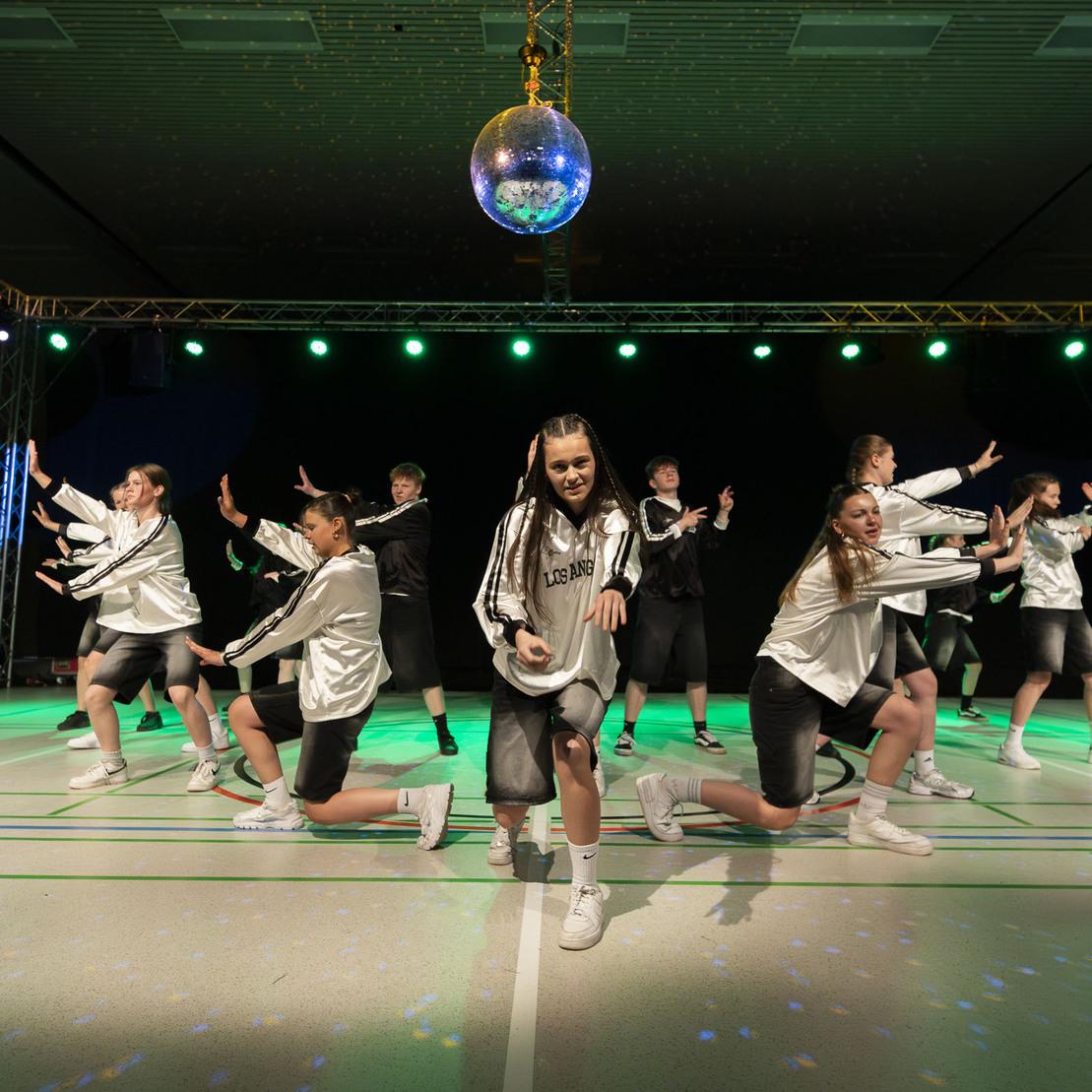 Tanzgruppe in Sportoutfits pr&auml;sentiert einen synchronen Auftritt auf einer beleuchteten B&uuml;hne mit Discokugel.