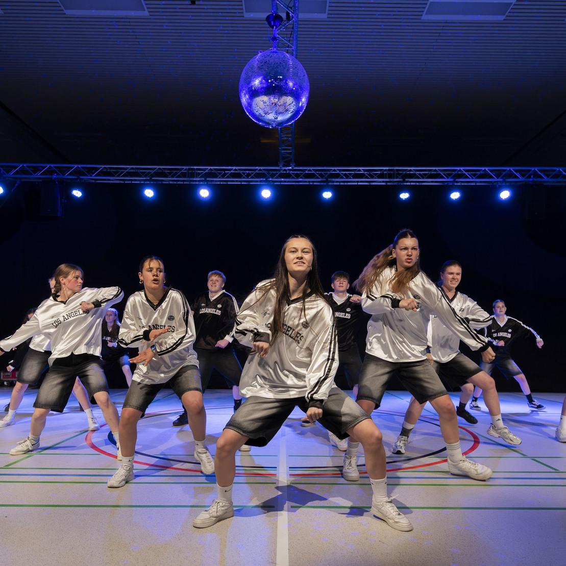 Tanzgruppe in silbernen Outfits f&uuml;hrt eine Choreografie unter einer Discokugel auf B&uuml;hne mit blauer Beleuchtung auf.