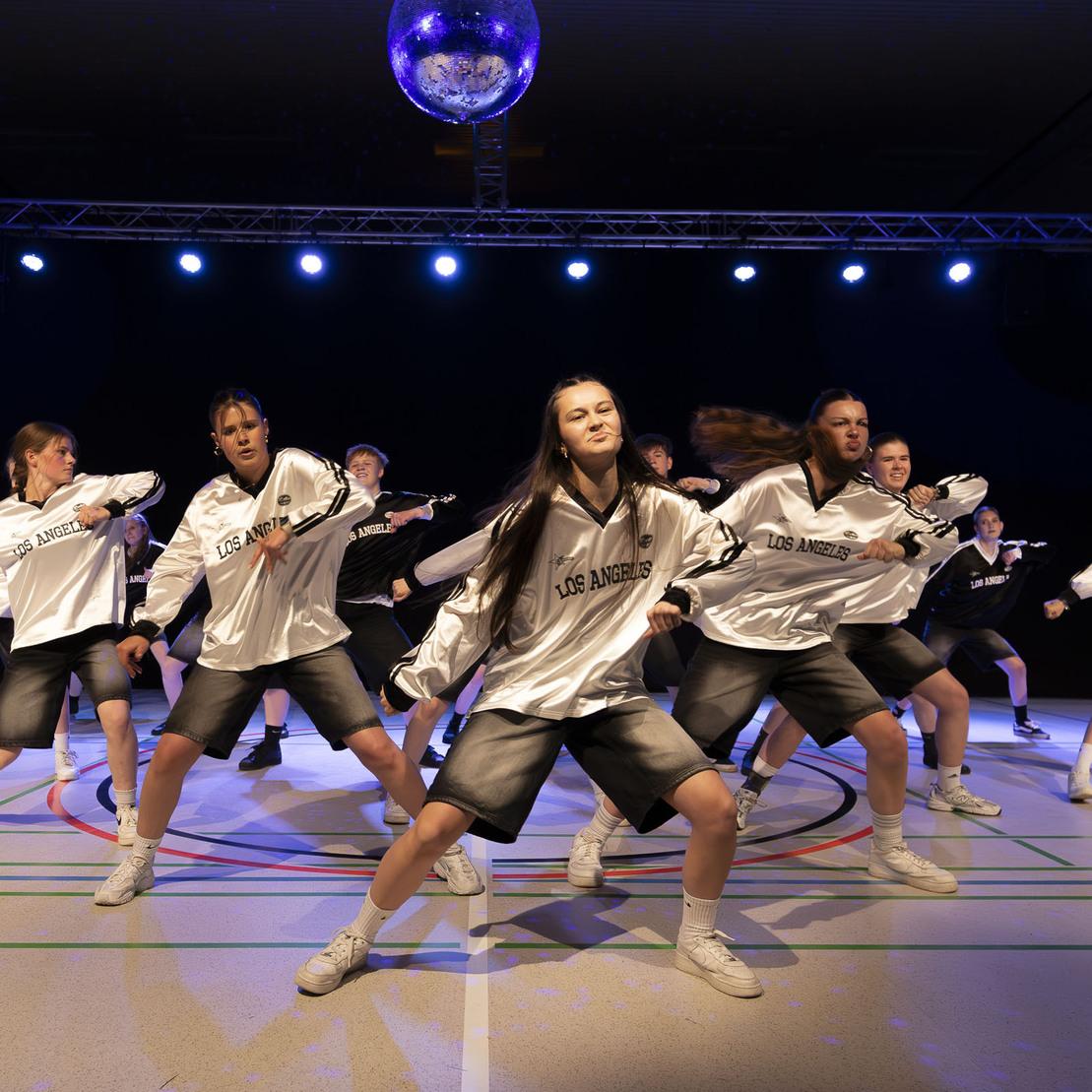 Tanzgruppe in wei&szlig;en Shirts und Shorts f&uuml;hrt eine Choreografie unter bunten Lichtern auf einer Sportfl&auml;che vor.