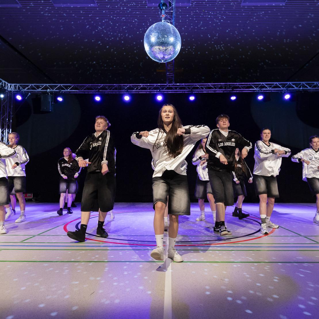 Tanzgruppe in Sportkleidung f&uuml;hrt eine Choreografie unter einer Discokugel in einer beleuchteten Halle auf.