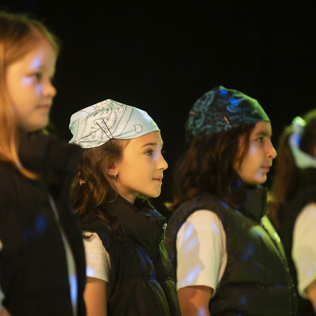 Vier Kinder in schwarzer Weste und bunten Kopfbedeckungen stehen nebeneinander auf einer B&uuml;hne mit farbigem Hintergrund.