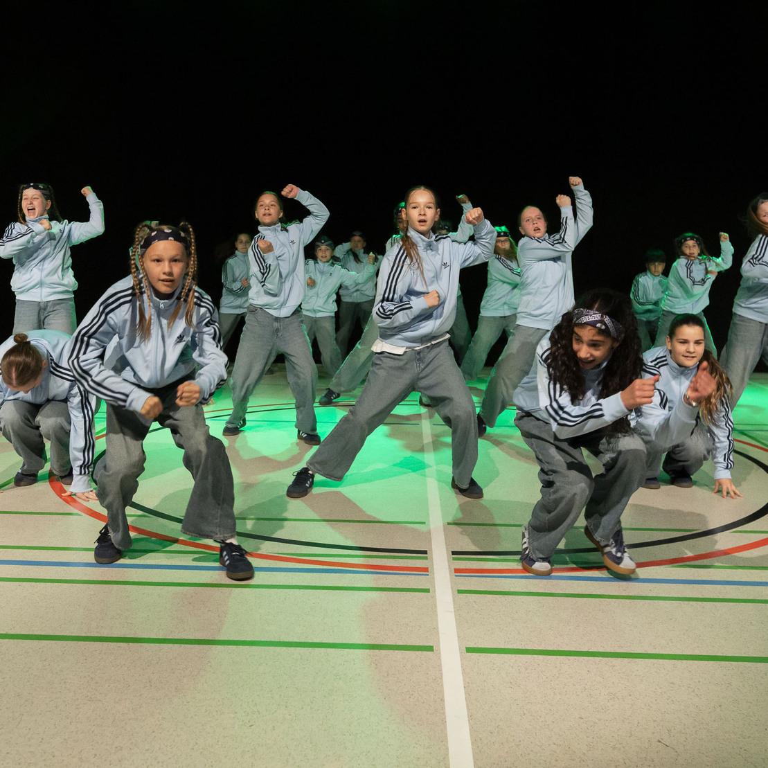 Tanzgruppe in grauen Trainingsanz&uuml;gen f&uuml;hrt eine dynamische Choreographie auf einer beleuchteten Sportfl&auml;che vor.