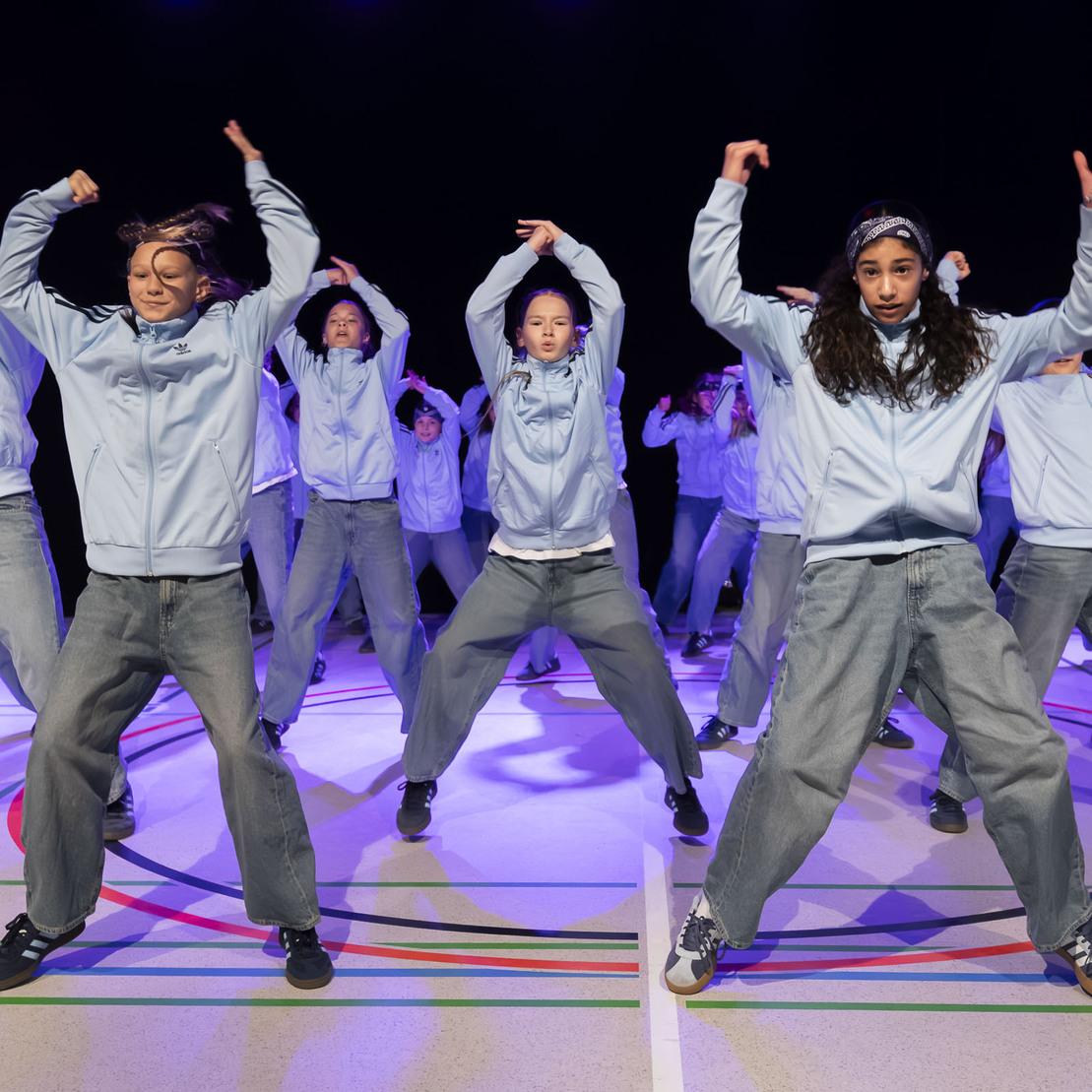 Tanzgruppe in blauen Sweatshirts und Jeans f&uuml;hrt Synchronbewegungen auf einer bunten B&uuml;hne aus.