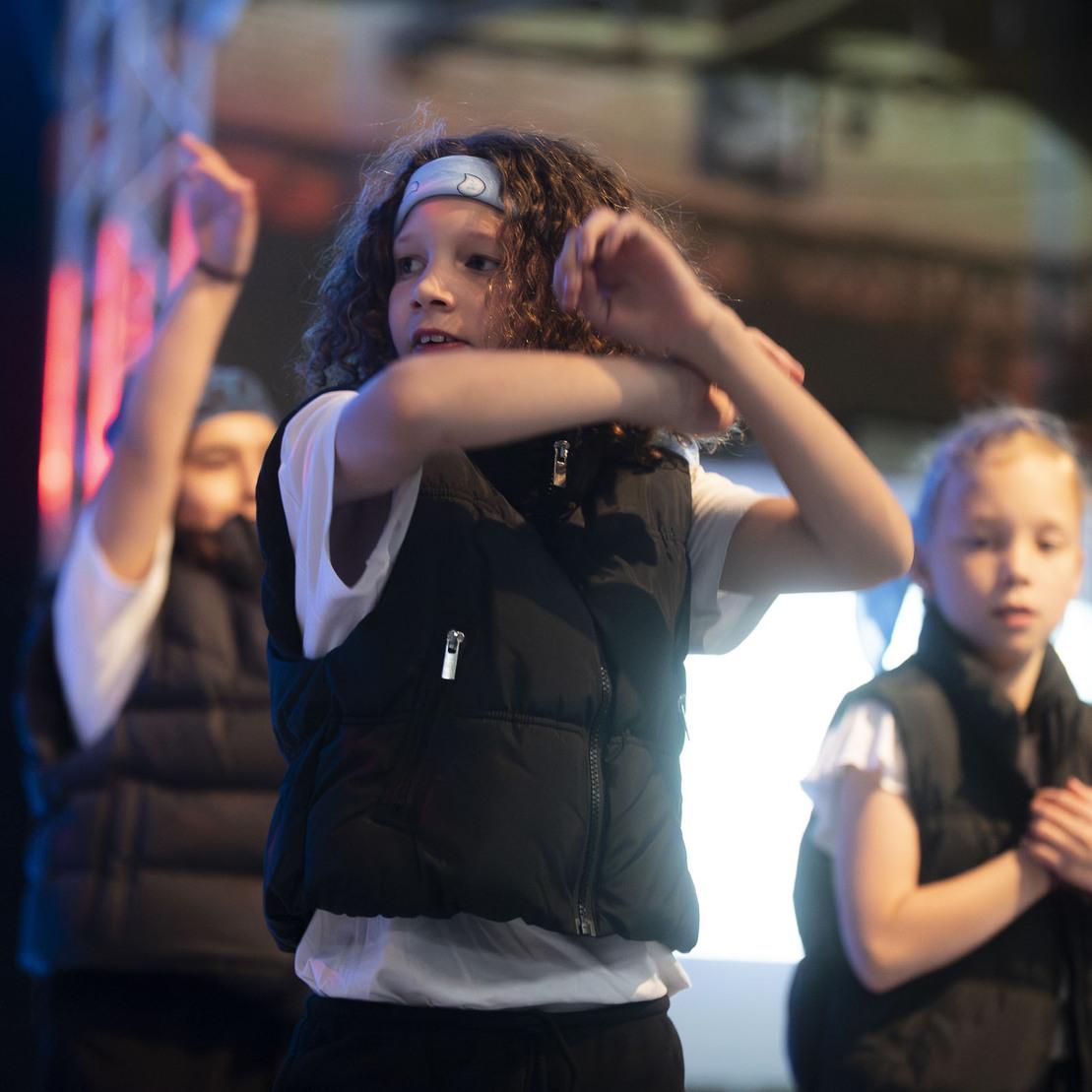 Tanzende Kinder in schwarzen Westen auf einer B&uuml;hne, beleuchtet von bunten Lichtern, zeigen eine choreografierte Performance.