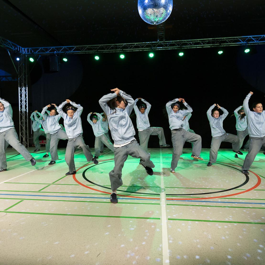 Tanzgruppe in grauen Outfits f&uuml;hrt synchronisierte Schritte auf einer beleuchteten B&uuml;hne aus.
