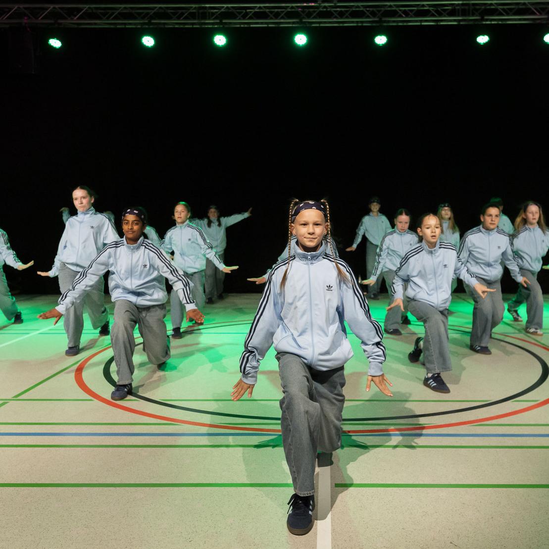 Tanzgruppe in blauen Trainingsanz&uuml;gen f&uuml;hrt eine Choreografie auf einer beleuchteten Sportfl&auml;che vor.