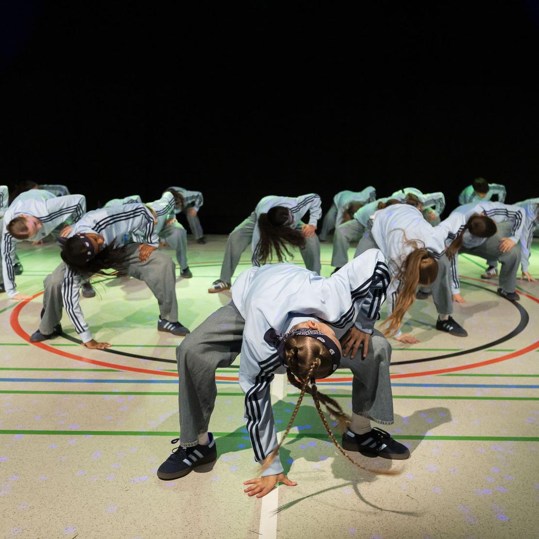 Gruppe von T&auml;nzern in Sportkleidung f&uuml;hrt synchronisierte Bewegungen auf einem beleuchteten Hallenboden aus.