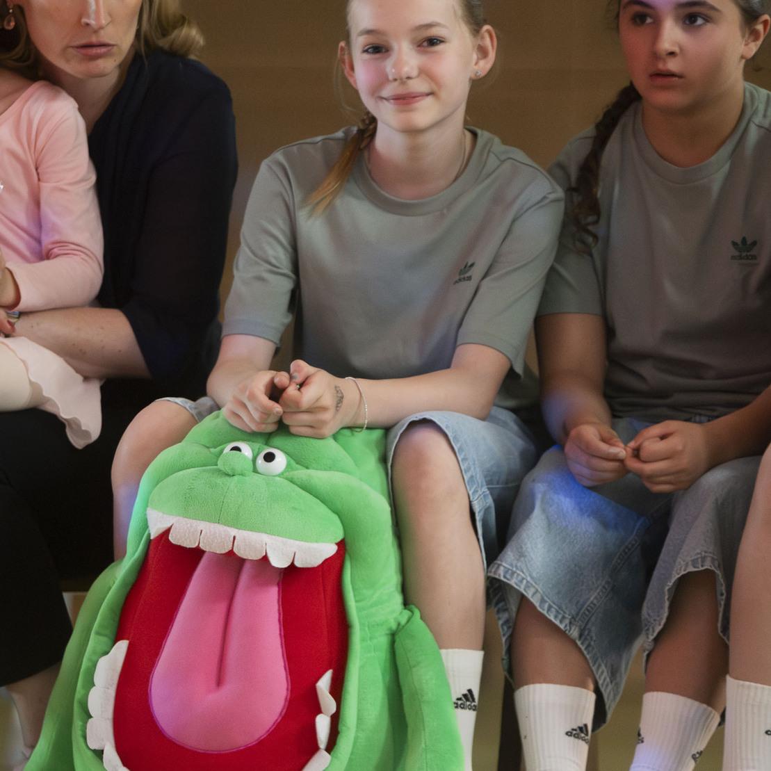 Eine Erwachsene und drei Kinder in Sportkleidung sitzen auf einer Bank, ein gr&uuml;nes Stofftier zeigt ein gro&szlig;es, offenes Maul.