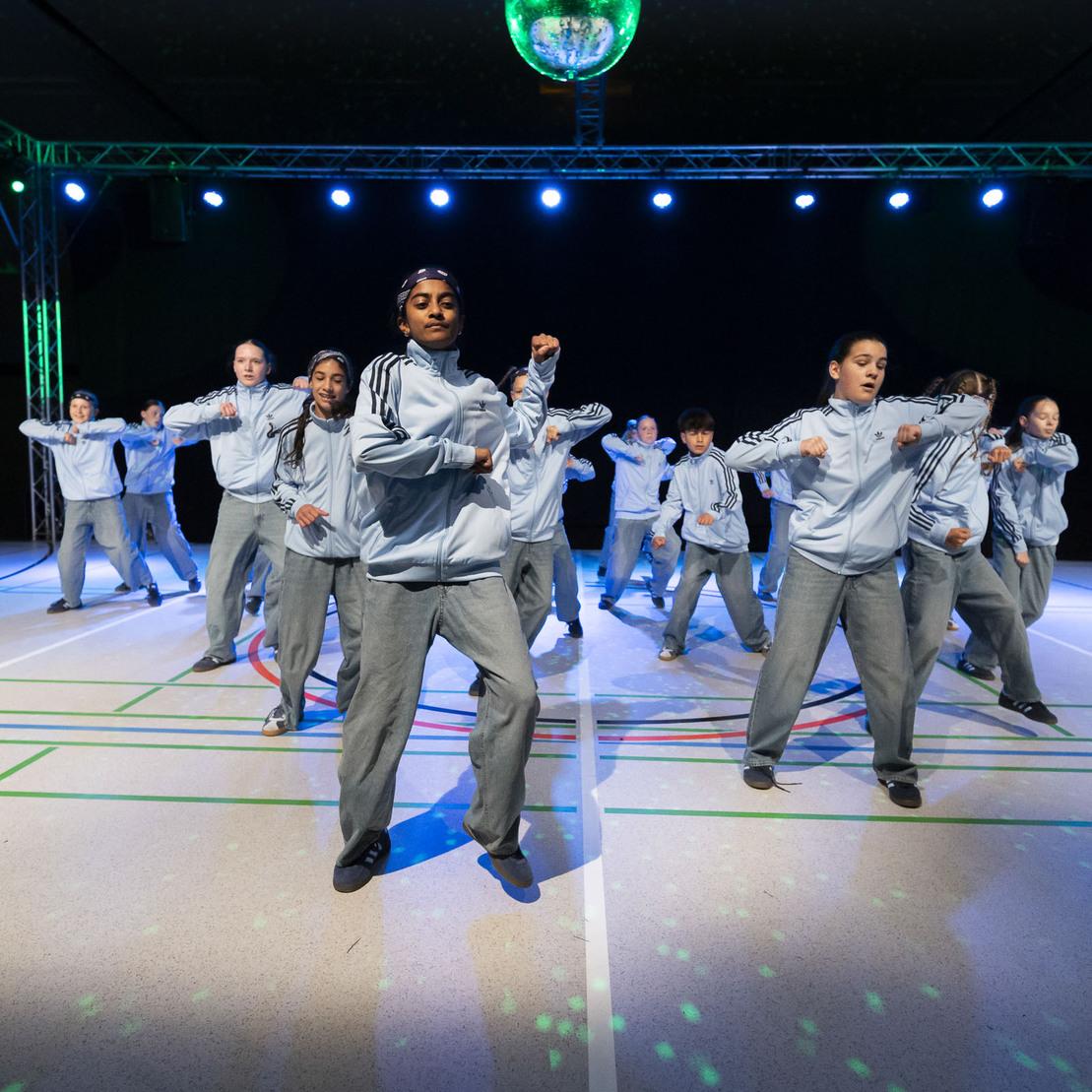Gruppe von T&auml;nzern in grauen Trainingsanz&uuml;gen f&uuml;hrt synchronisierte Moves auf einer beleuchteten Tanzfl&auml;che aus.