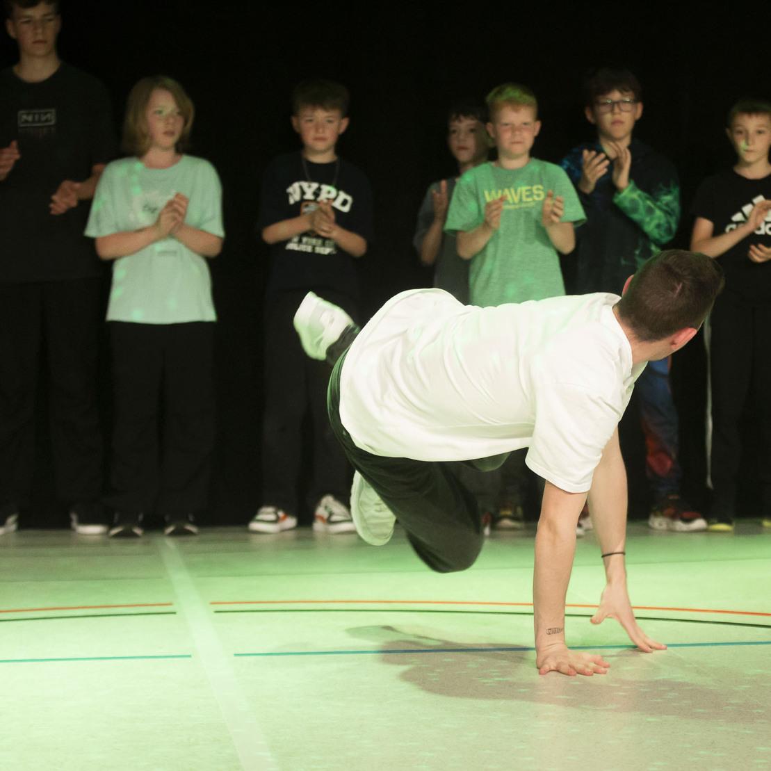 T&auml;nzer in wei&szlig;em T-Shirt f&uuml;hrt einen Breakdance-Move auf einer Sportfl&auml;che vor applaudierender Zuschauerschaft aus.