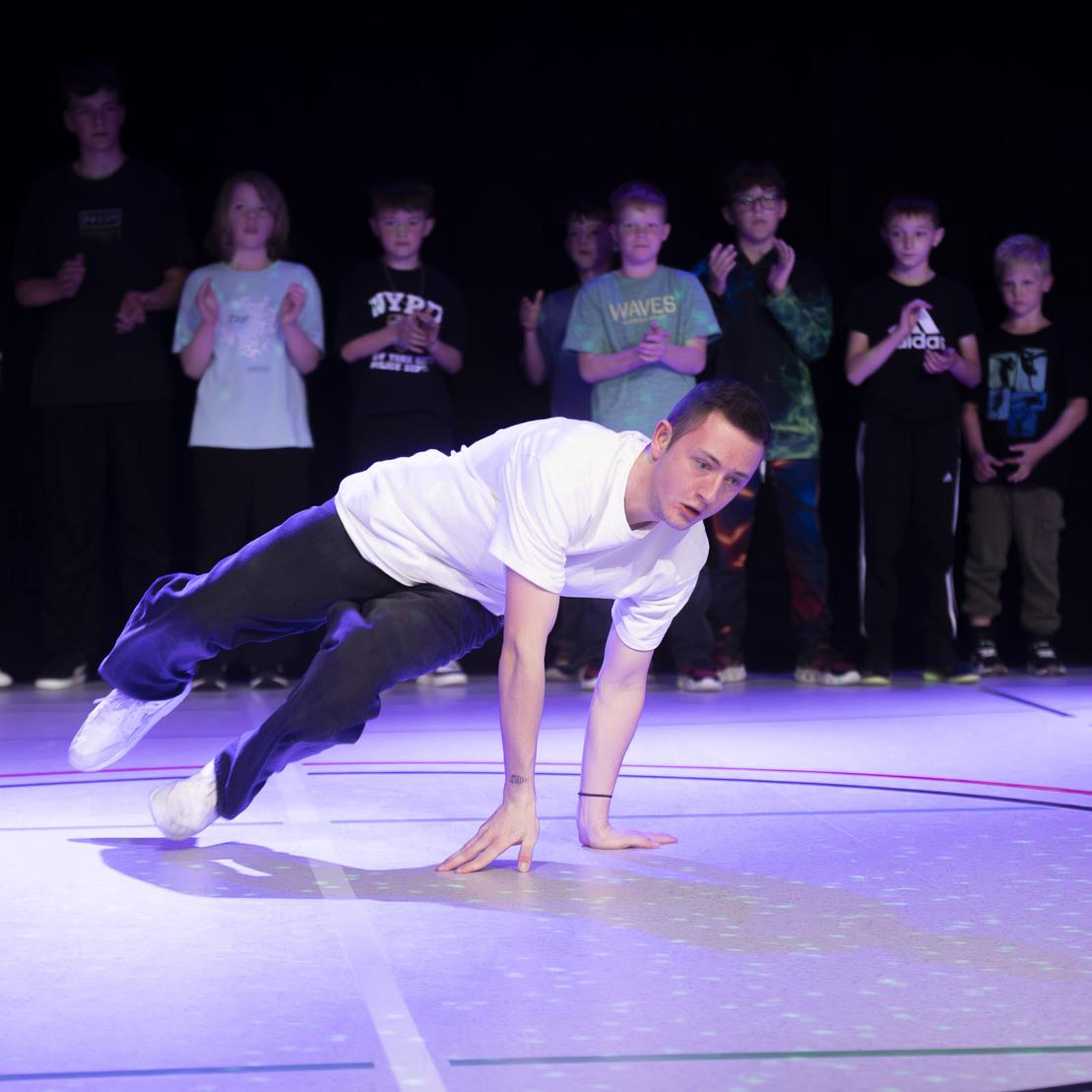 T&auml;nzer f&uuml;hrt eine Breakdance-Bewegung auf einer mit Lichtern beleuchteten B&uuml;hne vor applaudierendem Publikum aus.