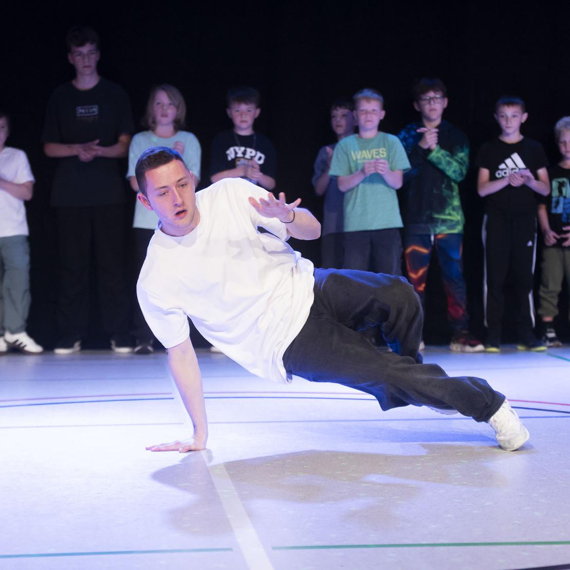 Breakdancer f&uuml;hrt eine Tanzbewegung auf einer B&uuml;hne aus, umgeben von zuschauenden Jugendlichen im Hintergrund.