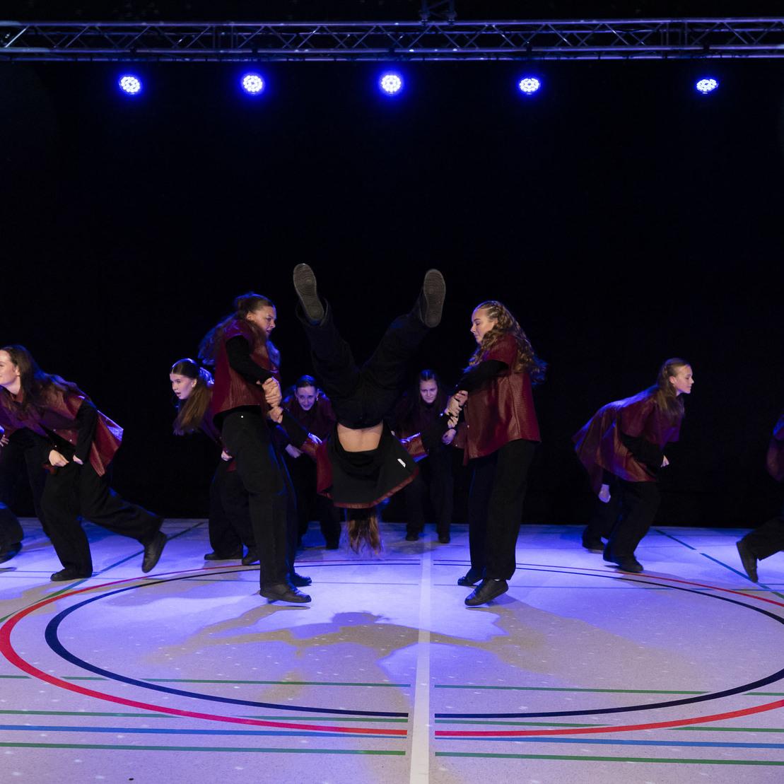 Tanzgruppe in modernen Outfits f&uuml;hrt akrobatische Bewegungen auf einer B&uuml;hne mit bunten Lichtern aus.