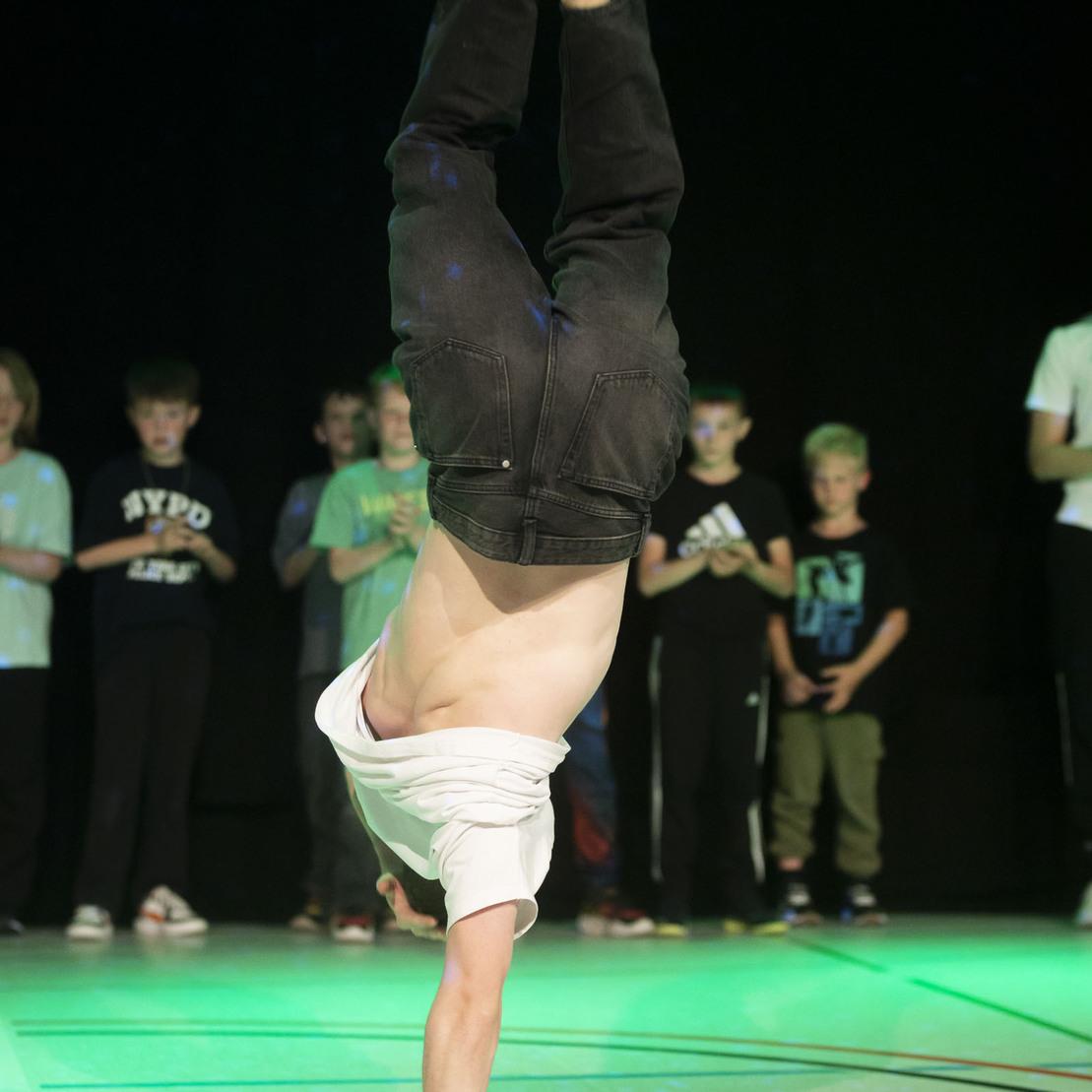 Breakdancer in Handstand-Position auf einer B&uuml;hne, umgeben von zuschauernden Kindern in der Hintergrundbeleuchtung.
