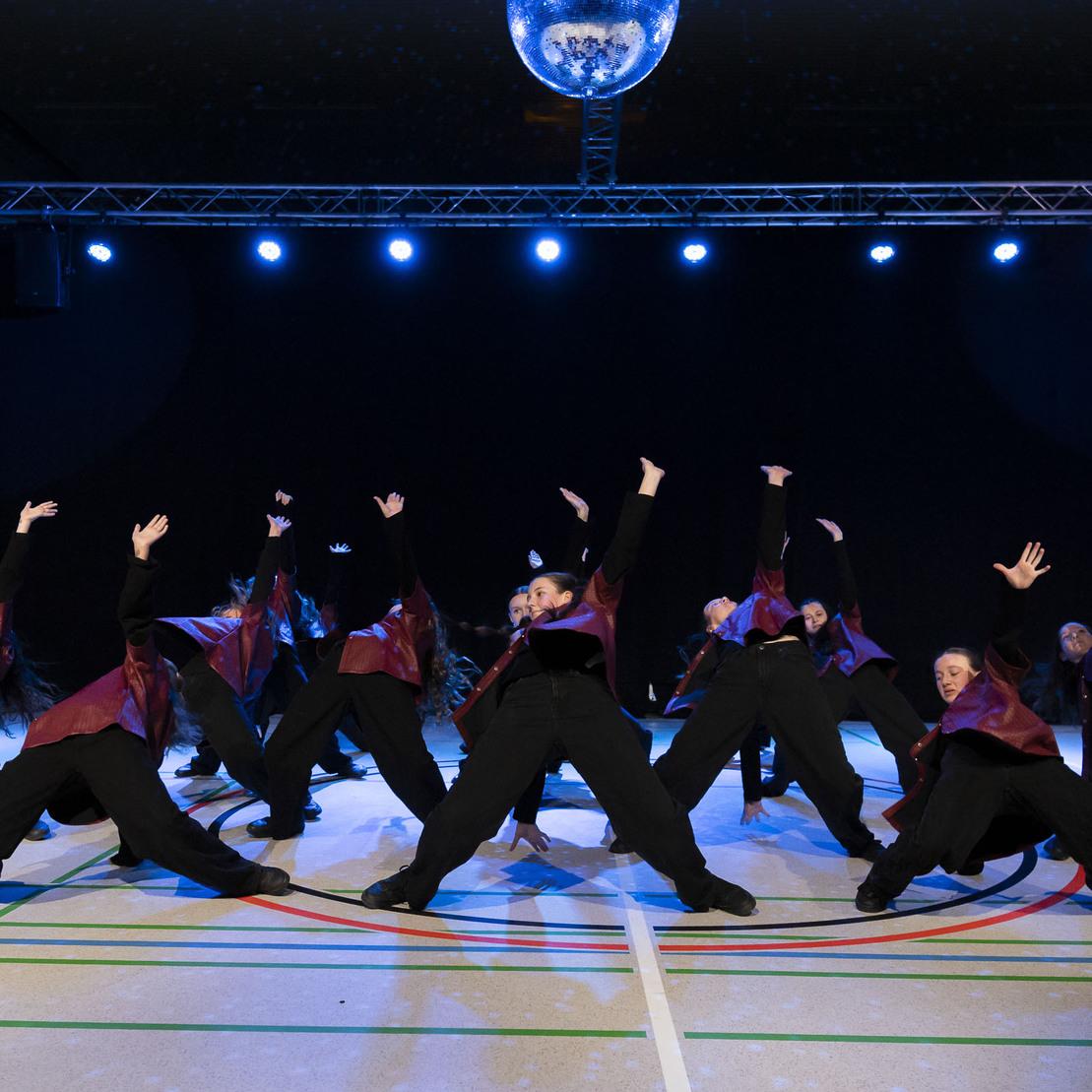Tanzgruppe in schwarzen Outfits mit roten Akzenten f&uuml;hrt synchronisierte Bewegungen unter einem Discokugel-Licht aus.