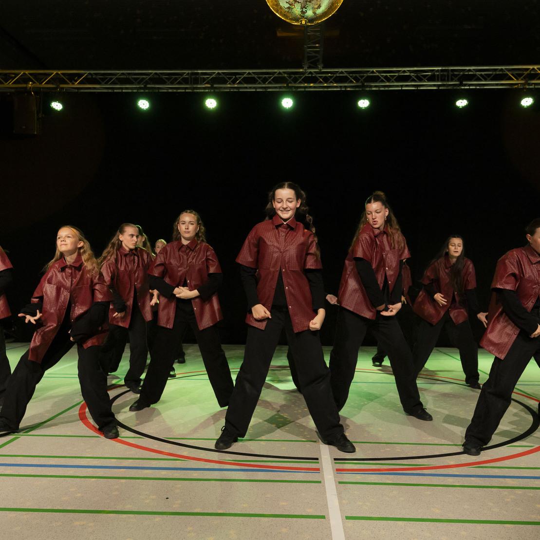 Gruppe von T&auml;nzerinnen in roten Outfits f&uuml;hrt synchronisierte Bewegungen auf einer beleuchteten B&uuml;hne aus.