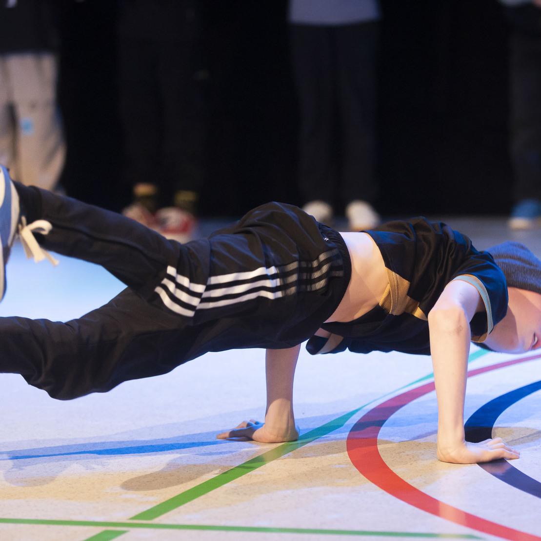 Jugendlicher in Sportkleidung f&uuml;hrt einen Breakdance-Move auf einem Tanzboden mit Linien durch.