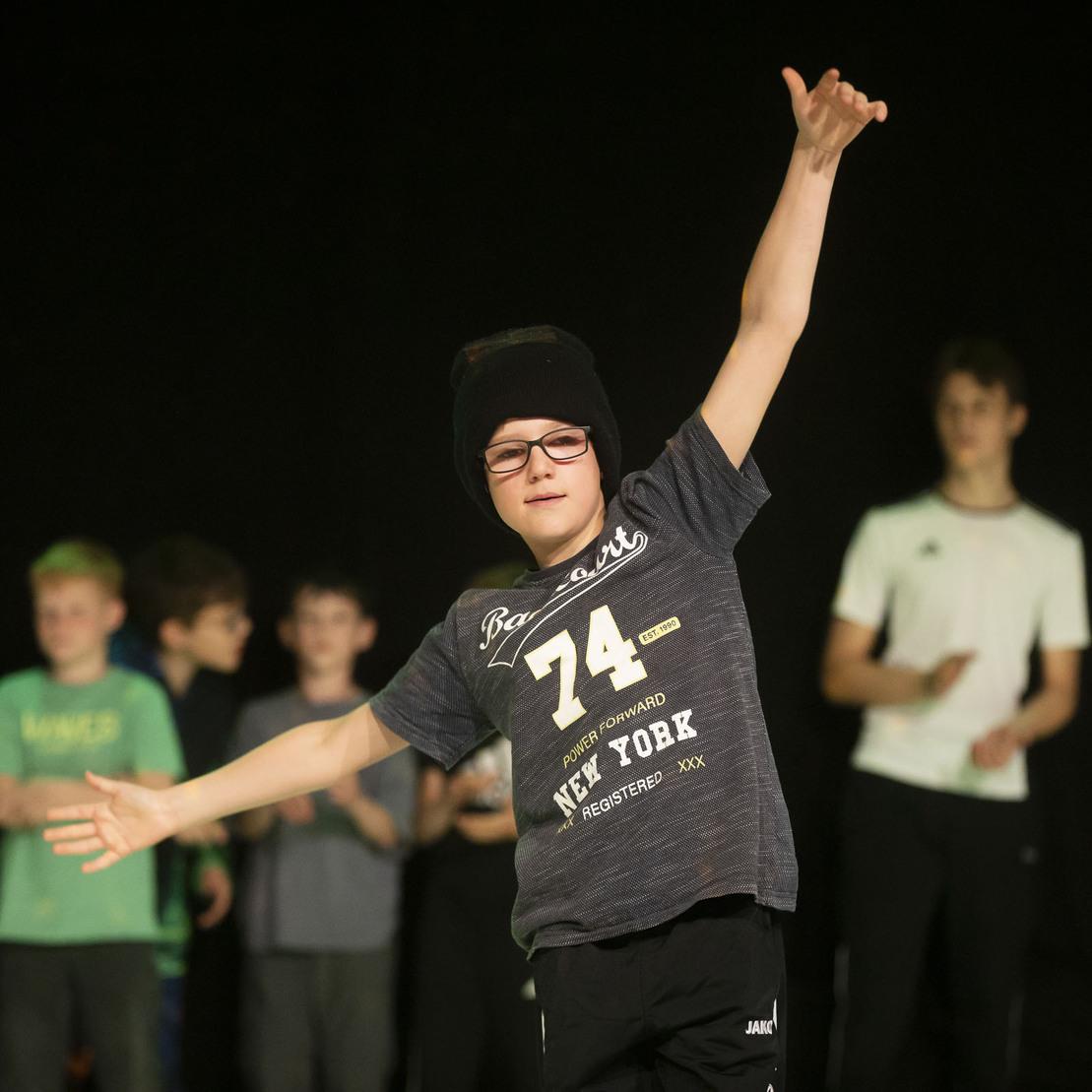 Ein Junge in einem schwarzen T-Shirt und M&uuml;tze tanzt vor einer Gruppe von Kindern auf einer B&uuml;hne mit dunklem Hintergrund.