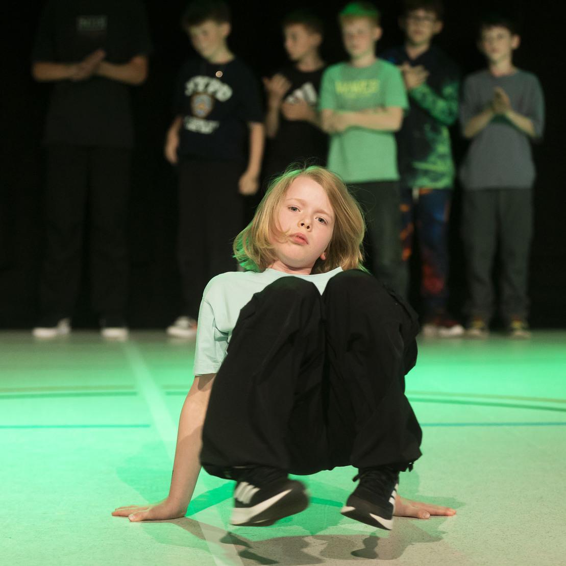 Ein Junge tanzt breakdance auf einem Sportboden, umgeben von applaudierenden Zuschauern im Hintergrund.