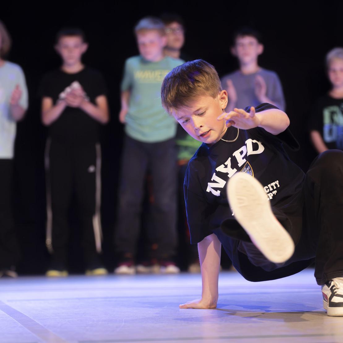 Ein Junge in schwarzem T-Shirt tanzt Breakdance auf einer B&uuml;hne, w&auml;hrend eine Gruppe Kinder applaudiert.
