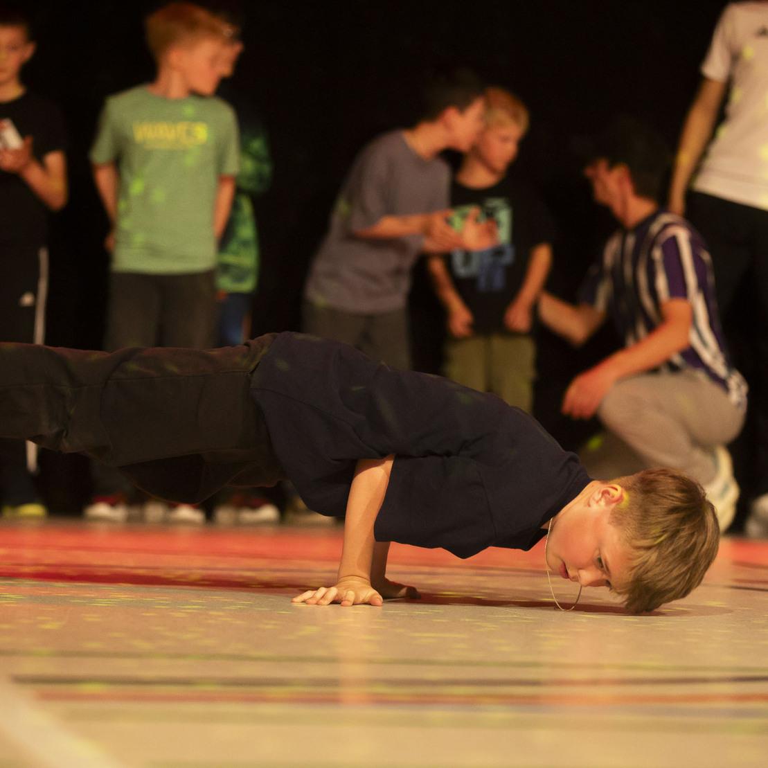 Ein Junge macht einen Handstand auf einer Sportfl&auml;che, w&auml;hrend andere Kinder im Hintergrund zuschauen.