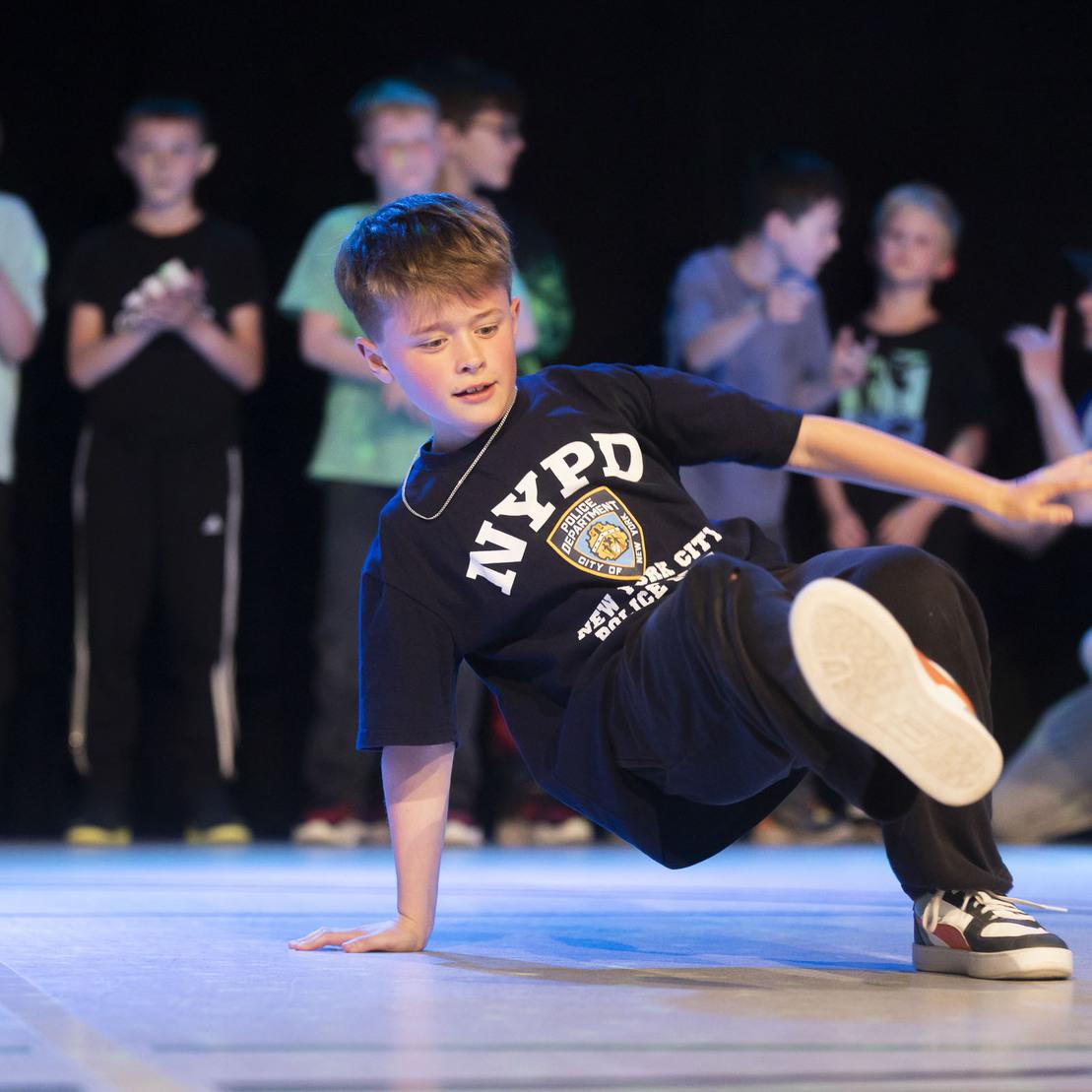 Ein Junge tanzt Breakdance auf der B&uuml;hne, umgeben von applaudierenden Jugendlichen im Hintergrund.