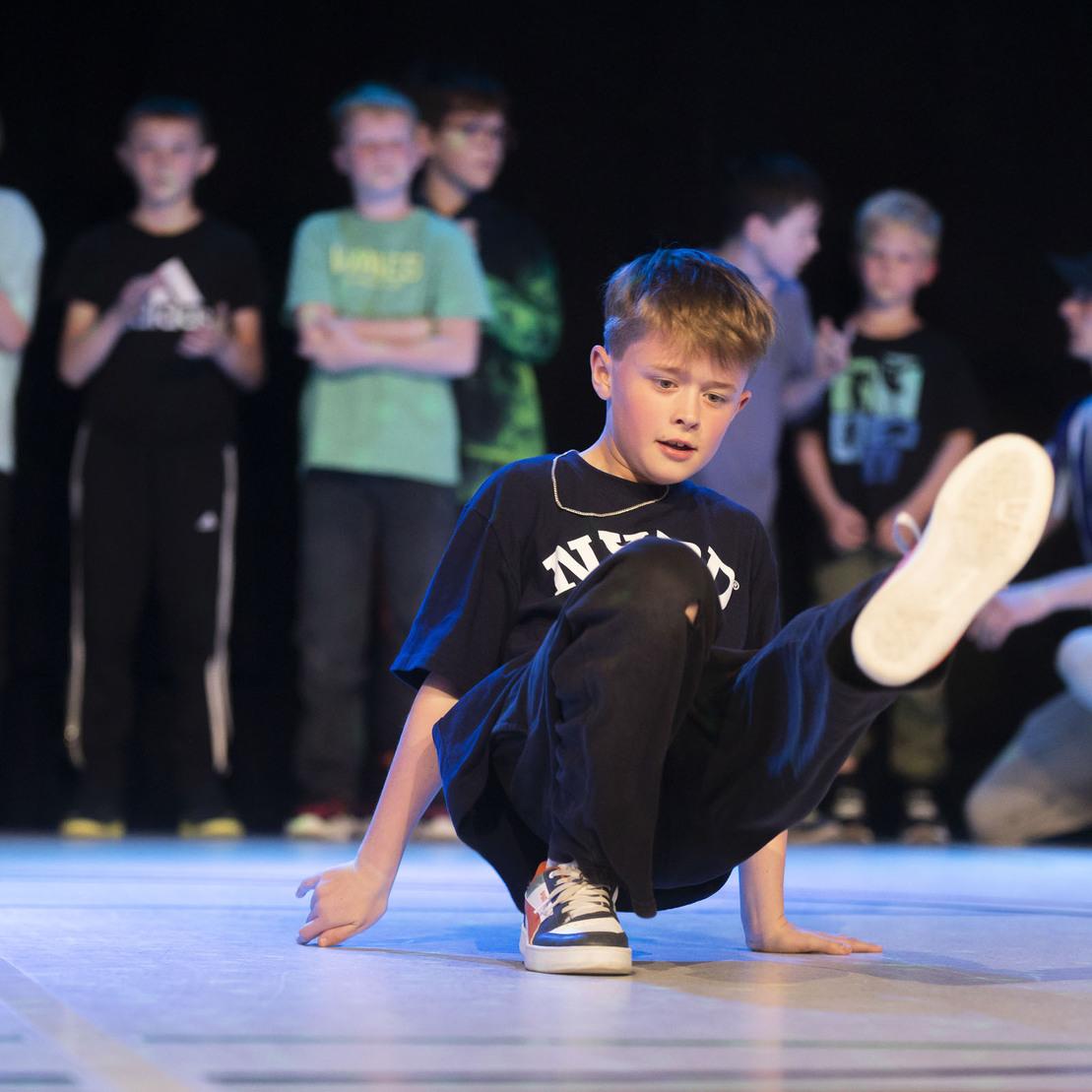 Ein Junge tanzt Breakdance auf einer B&uuml;hne, w&auml;hrend eine Gruppe Zuschauer im Hintergrund zuschaut.