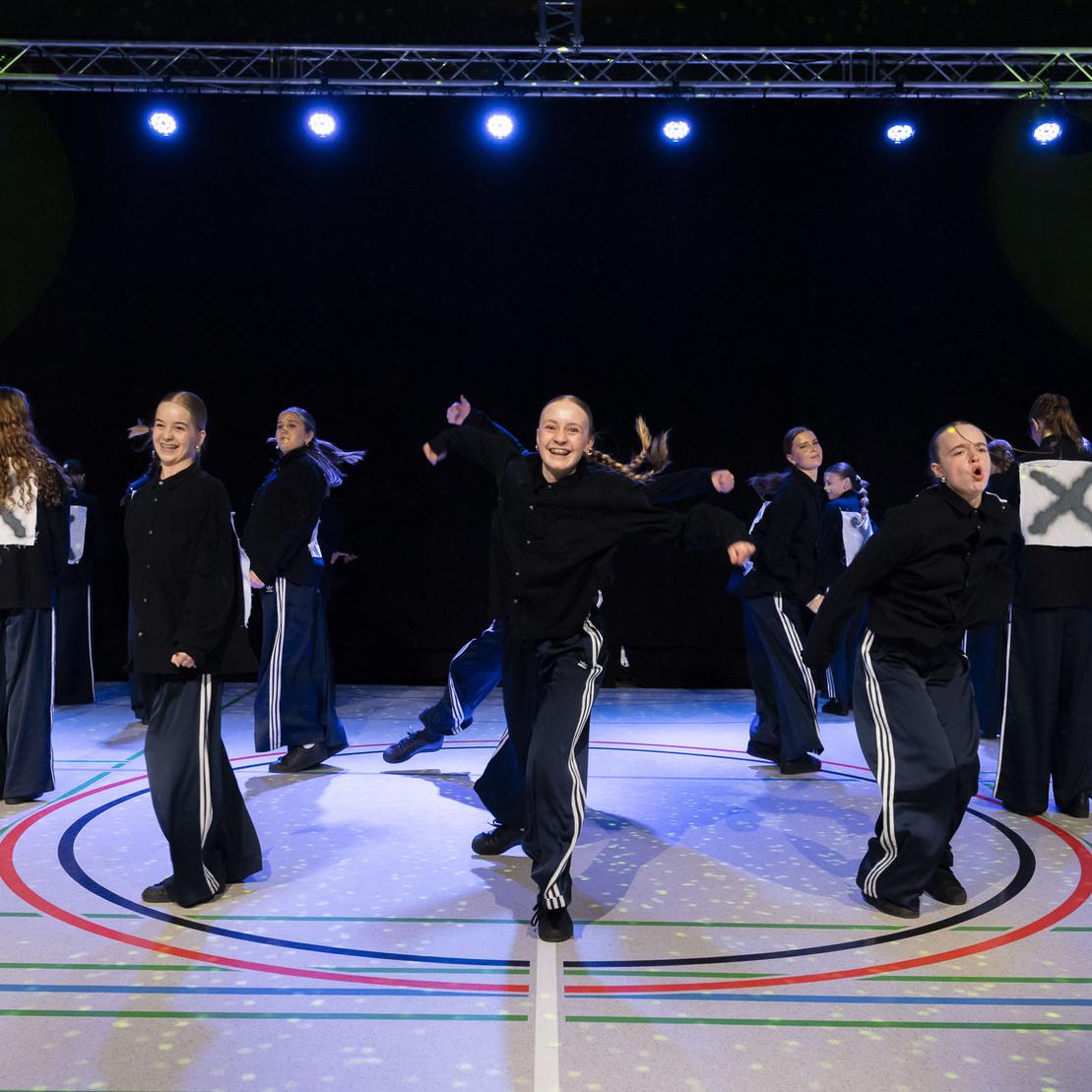 Tanzgruppe in schwarzen Outfits mit X-Symbolen, die eine auff&auml;llige Choreografie auf einer beleuchteten B&uuml;hne pr&auml;sentiert.