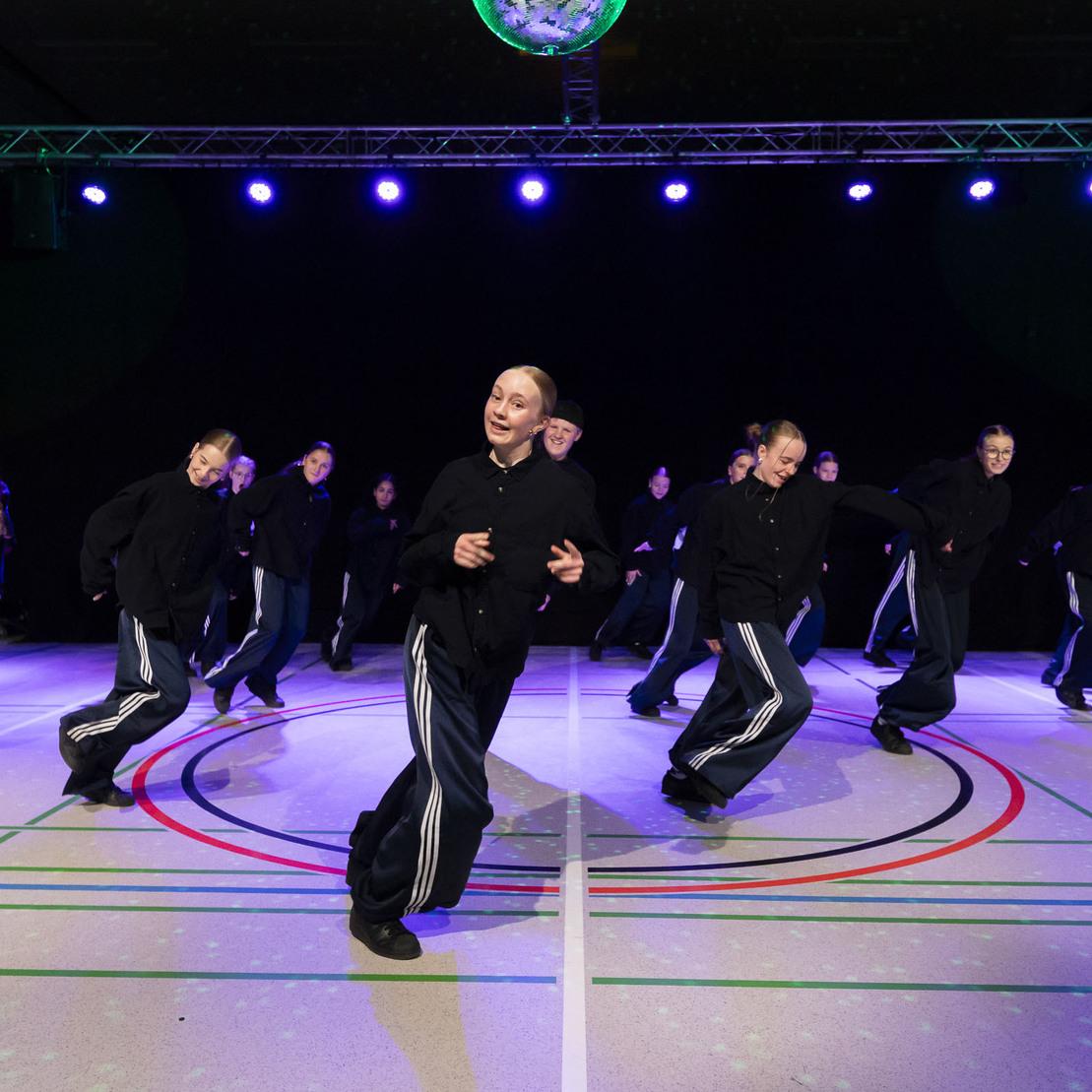 Tanzgruppe in schwarzen Outfits, die synchron auf einer beleuchteten Tanzfl&auml;che mit Spiegelkugel performt.