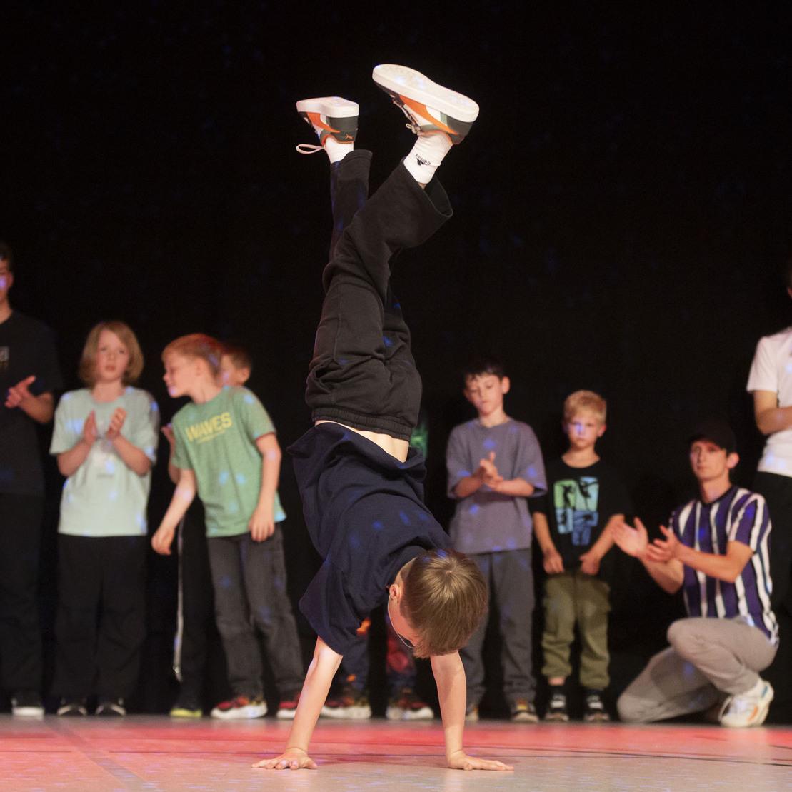Junge macht einen Handstand auf einer B&uuml;hne, w&auml;hrend eine Gruppe von Zuschauern applaudiert.