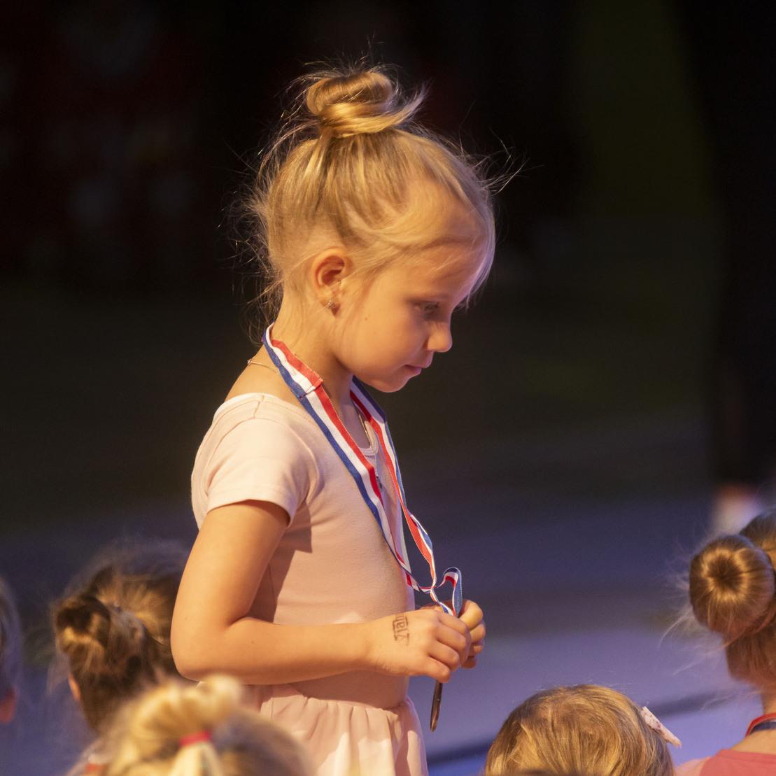 M&auml;dchen mit Medaille in rosa Kleidung steht auf einer B&uuml;hne, umgeben von anderen Kindern mit Hochsteckfrisuren.