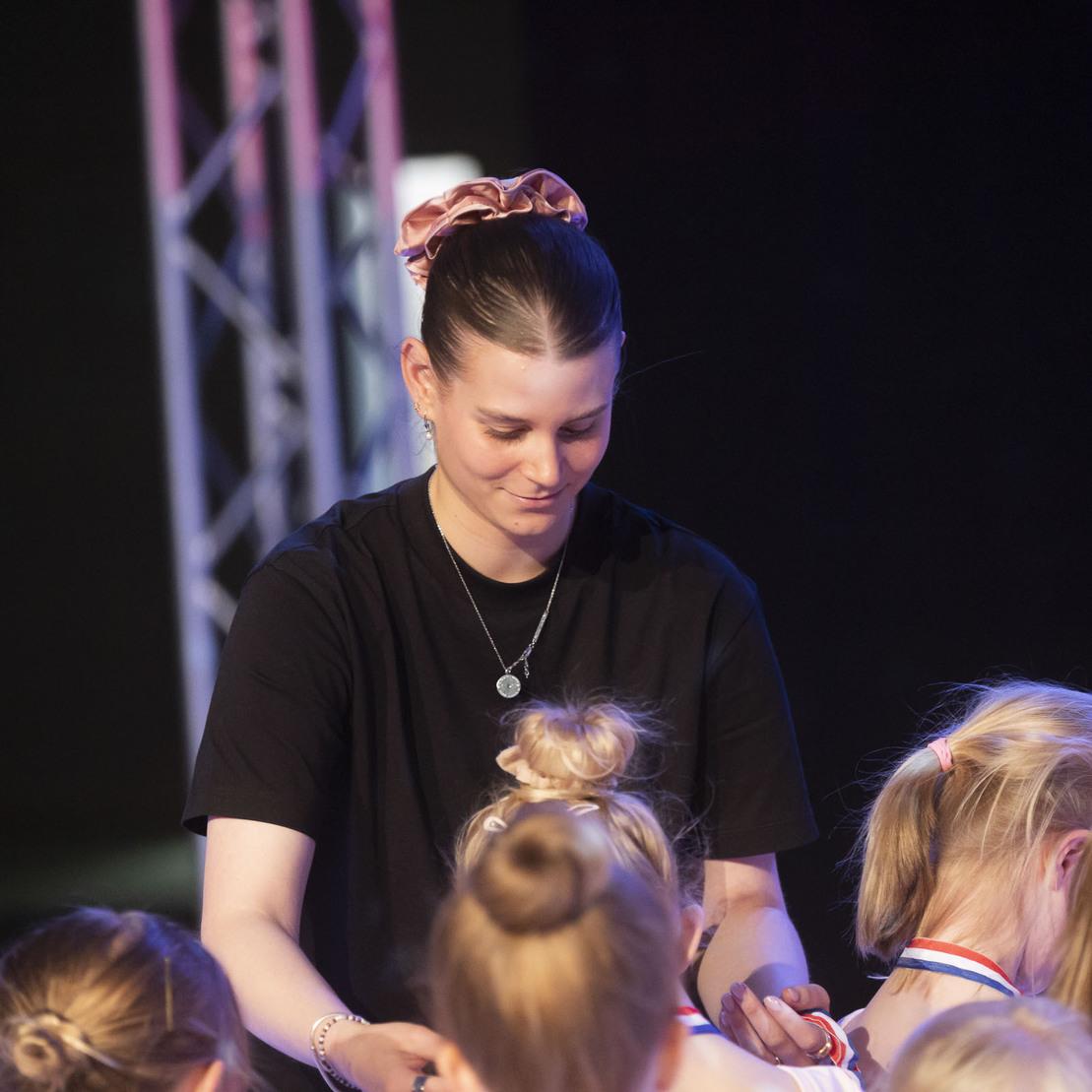 Eine Trainerin verteilt Medaillen an junge Teilnehmerinnen einer Sportveranstaltung. Bunte Lichter im Hintergrund.